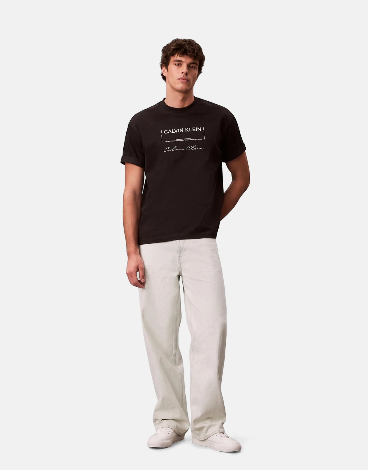 A male model stands wearing a black Calvin Klein 16s April Graphic Seasonal Fashion t-shirt with light jeans and white