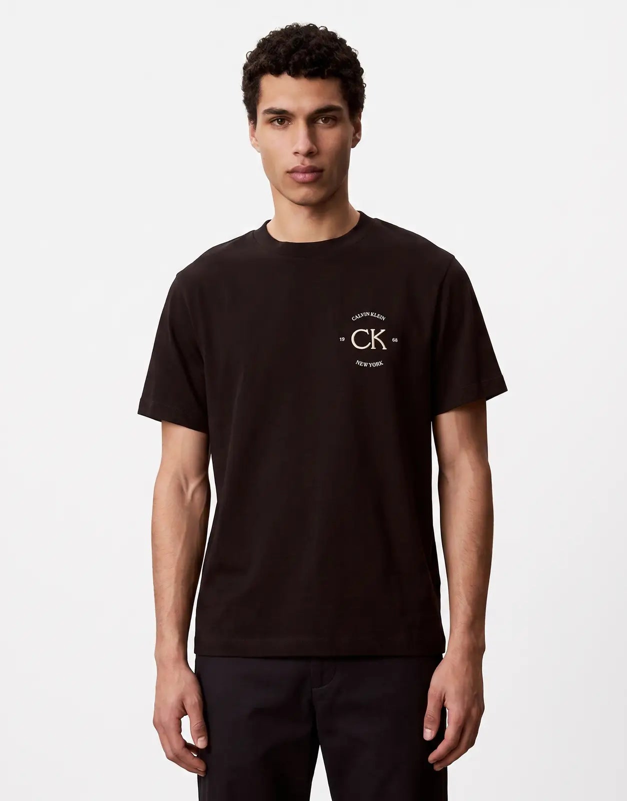 A young man models a black Calvin Klein 20s April Graphic Seasonal Fashion T-shirt with a white logo on a plain background.