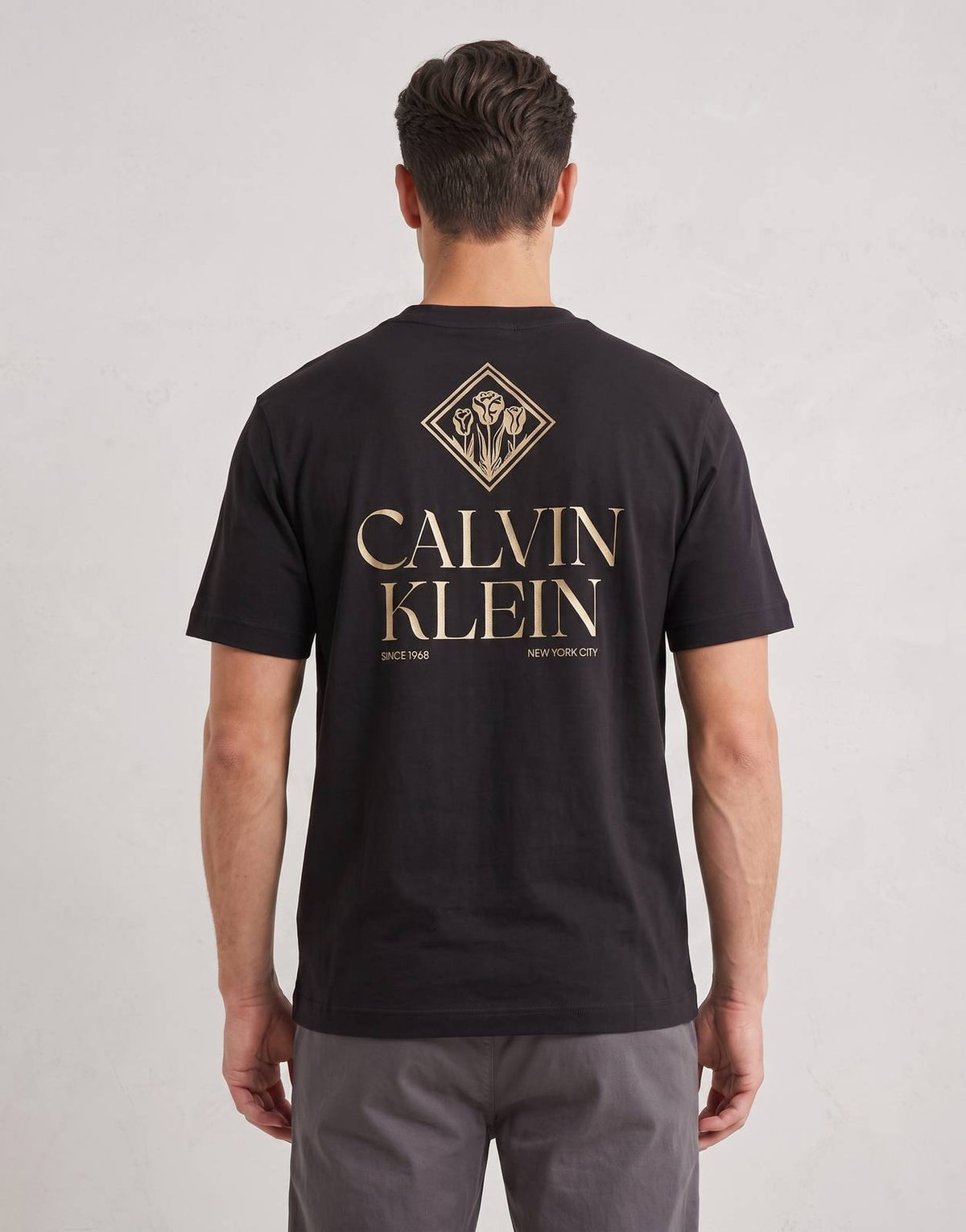 Men's Calvin Klein 20s Roses Graphic T-shirt in black, featuring a gold graphic logo on the back, modeled for e-commerce.