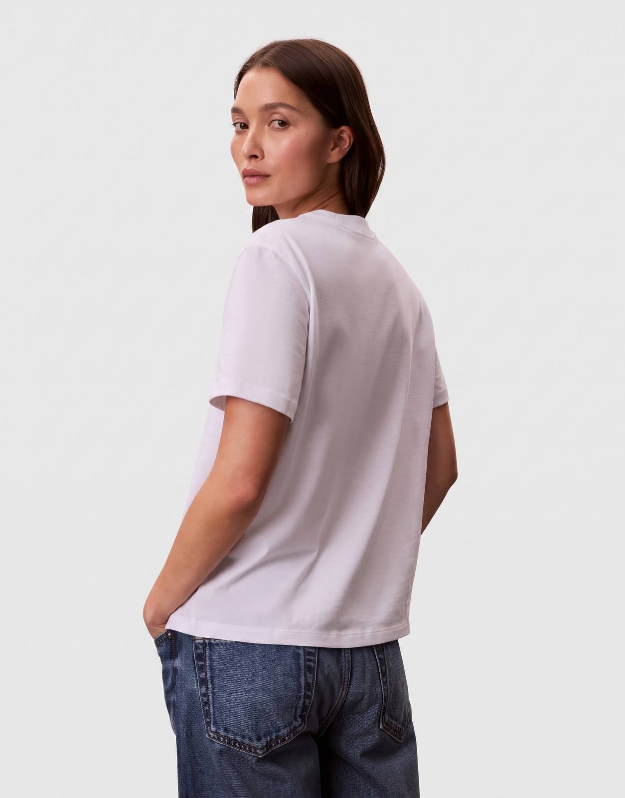 A female model wearing a Calvin Klein Classic Graphic White T-Shirt, shown from the back, paired with blue jeans. The model