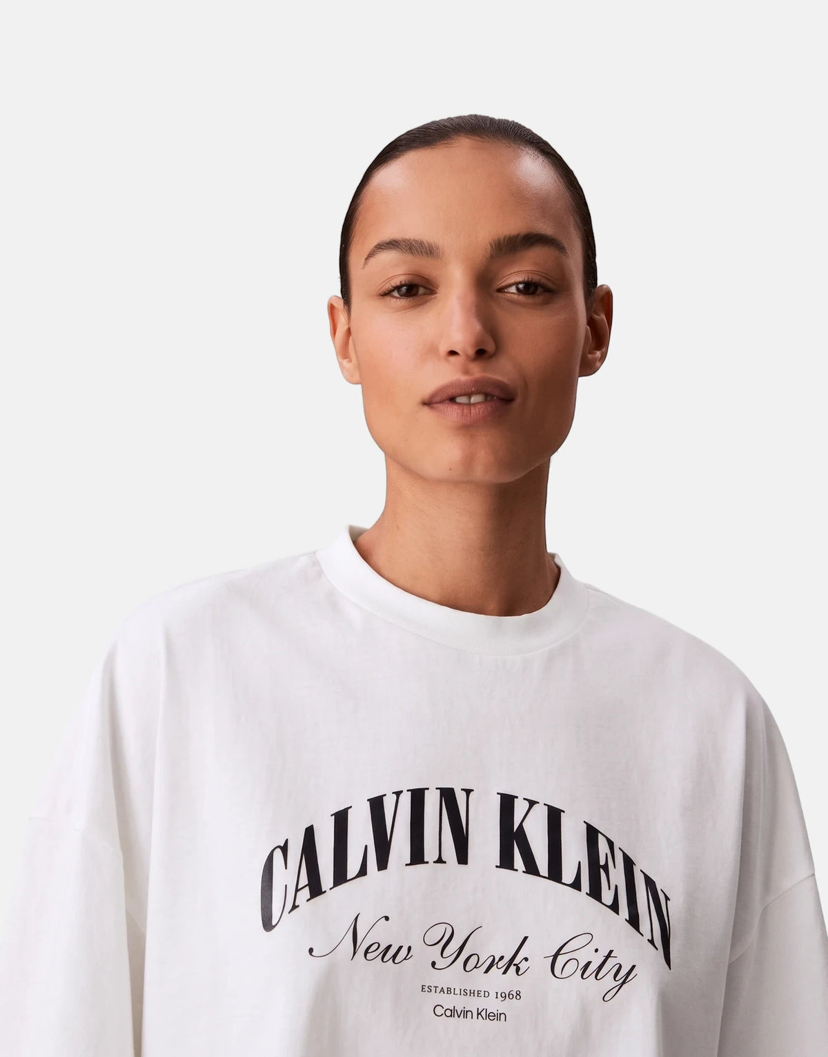 A female model wearing a white Calvin Klein Cropped Boxy Heritage Crest Logo T-Shirt with a black logo.