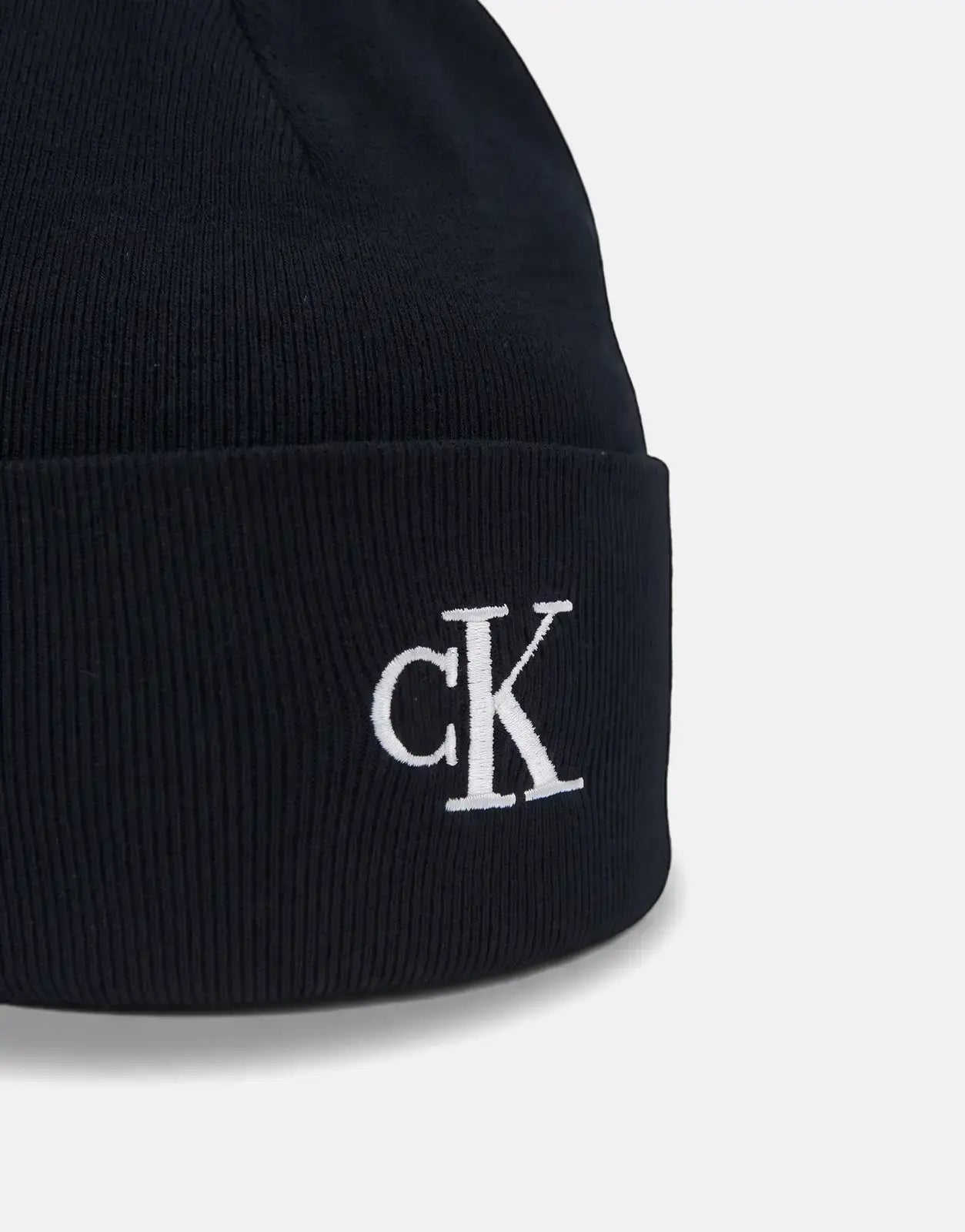 Close-up of a black Calvin Klein Fine Rib Beanie with a white embroidered CK logo on the front, resting on a surface.