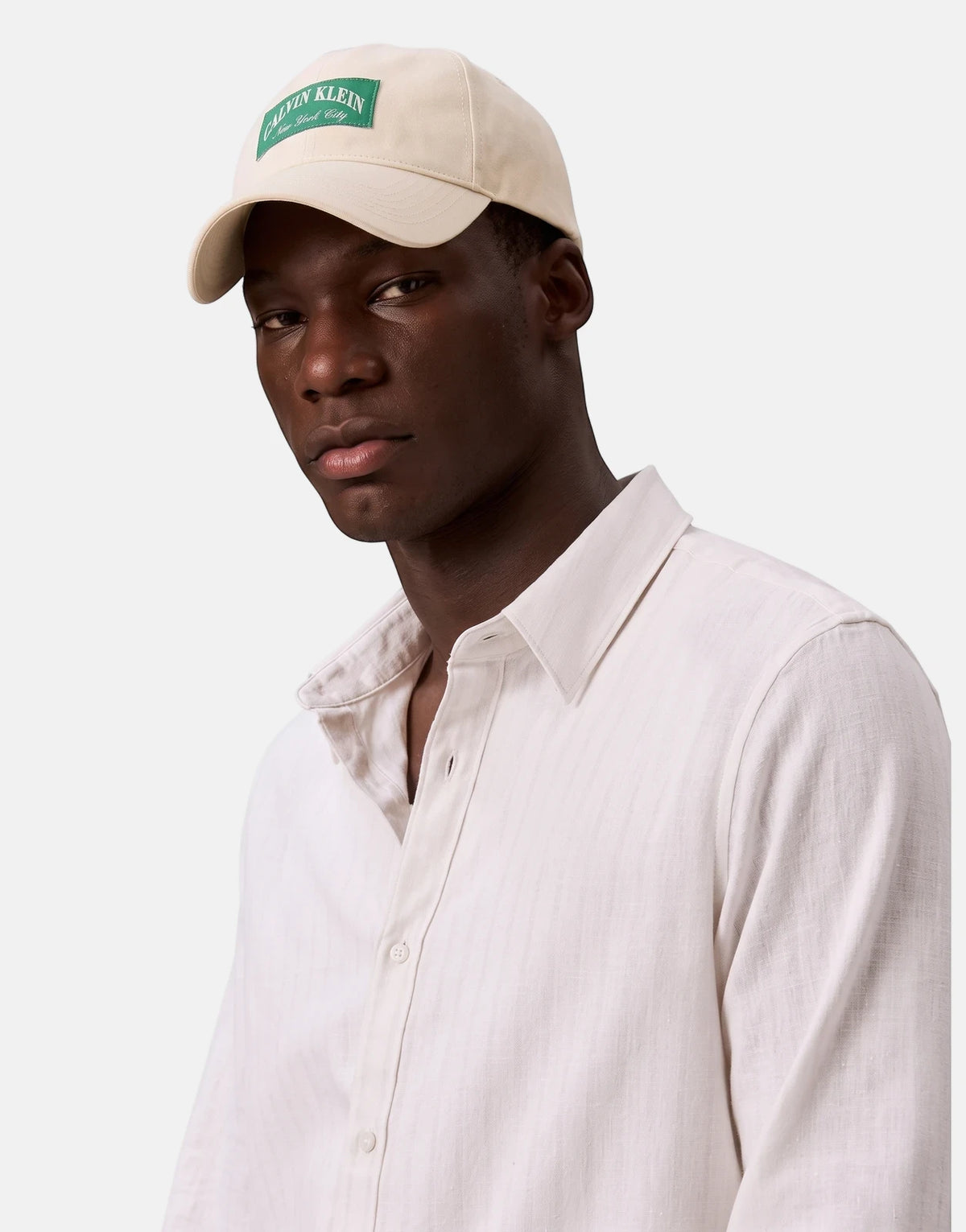 Male model wearing an off-white Calvin Klein Heritage Crest Pitch Cap with a green logo and a light-colored button-up shirt.
