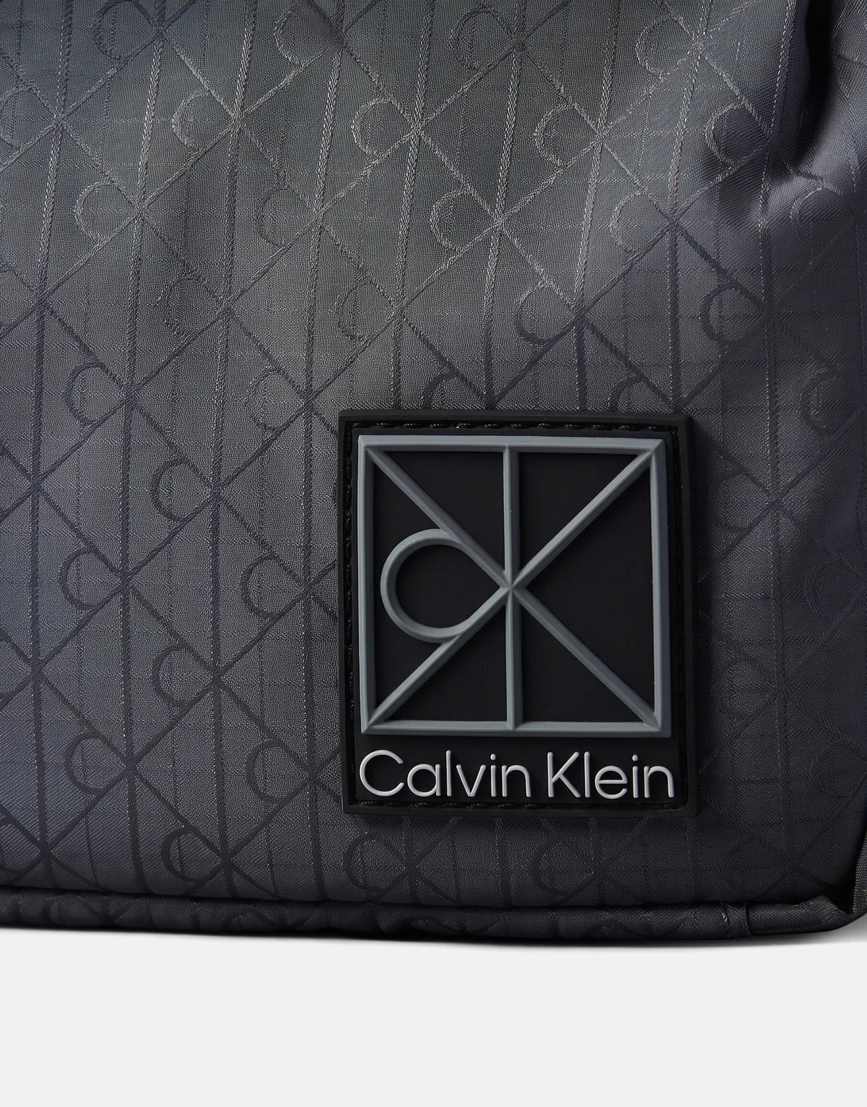 Close-up of a Calvin Klein Utility Emblem Reporter Bag in blue grey, showcasing the brand's logo patch and patterned fabric.