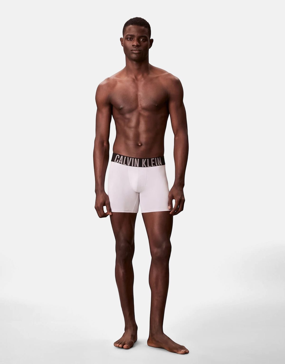 Male model showcasing Calvin Klein white boxer briefs with a black branded waistband, full body shot.