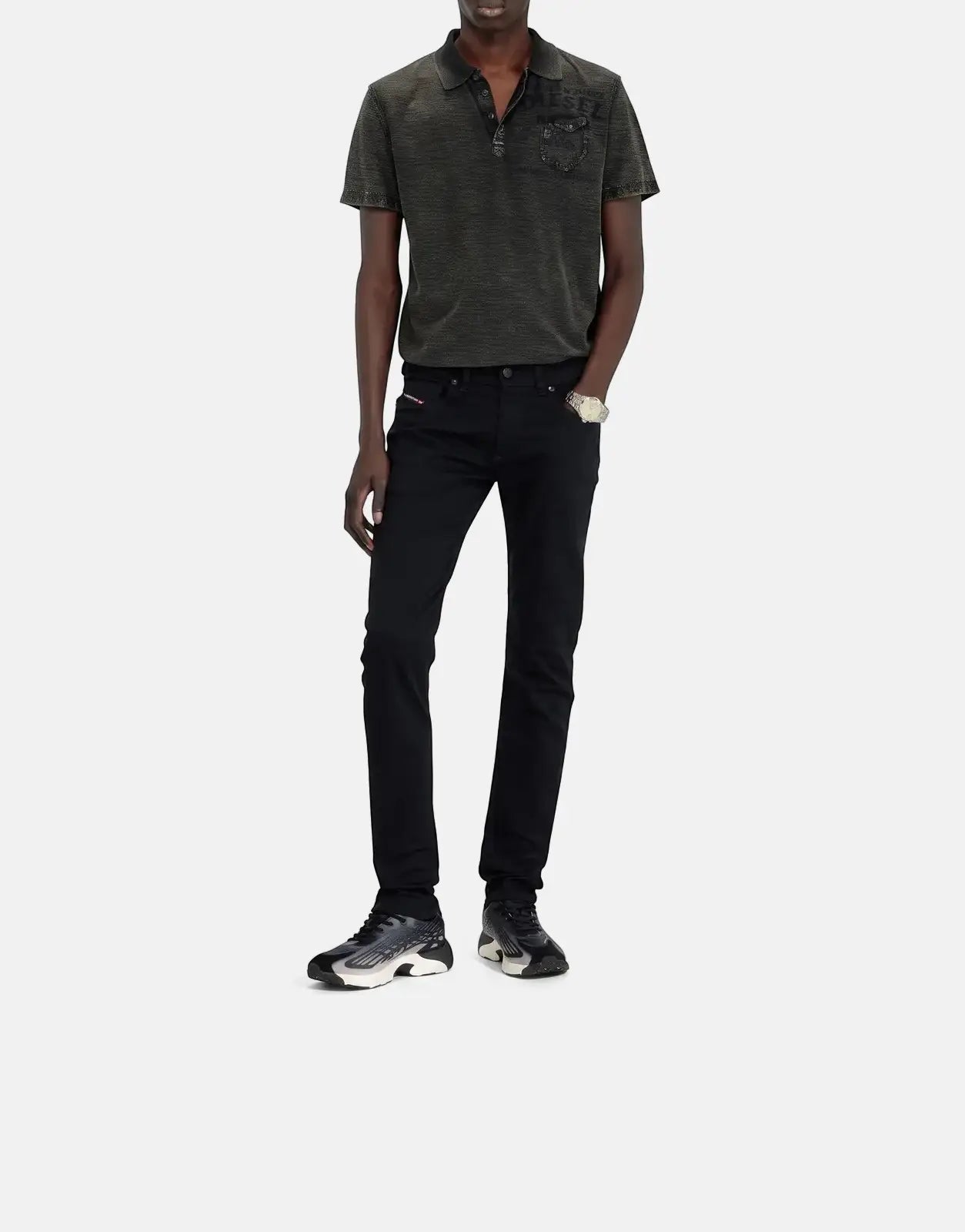 Full body shot of a male model wearing black Diesel 1979 Sleenker skinny jeans, a dark polo shirt, sunglasses, and sneakers.