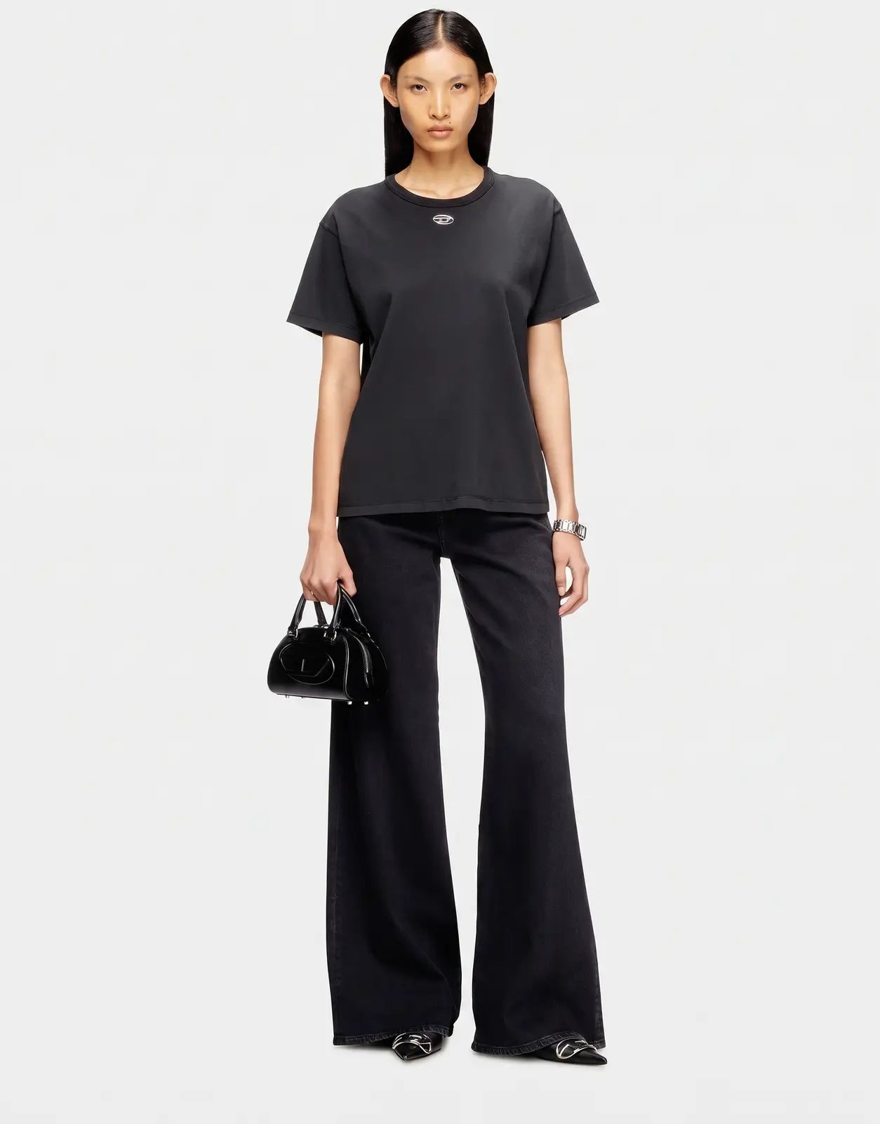 A female model wears a black regular fit Diesel T-Normy OD S1 T-shirt with black flared jeans, a black handbag, and black