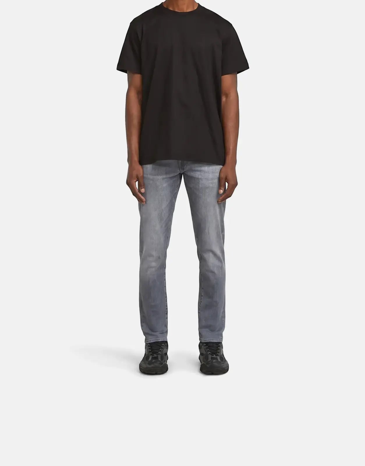 Full-body image of a man wearing G-Star RAW 3301 Sun Faded Glacier Grey jeans with a black t-shirt and sneakers, standing on