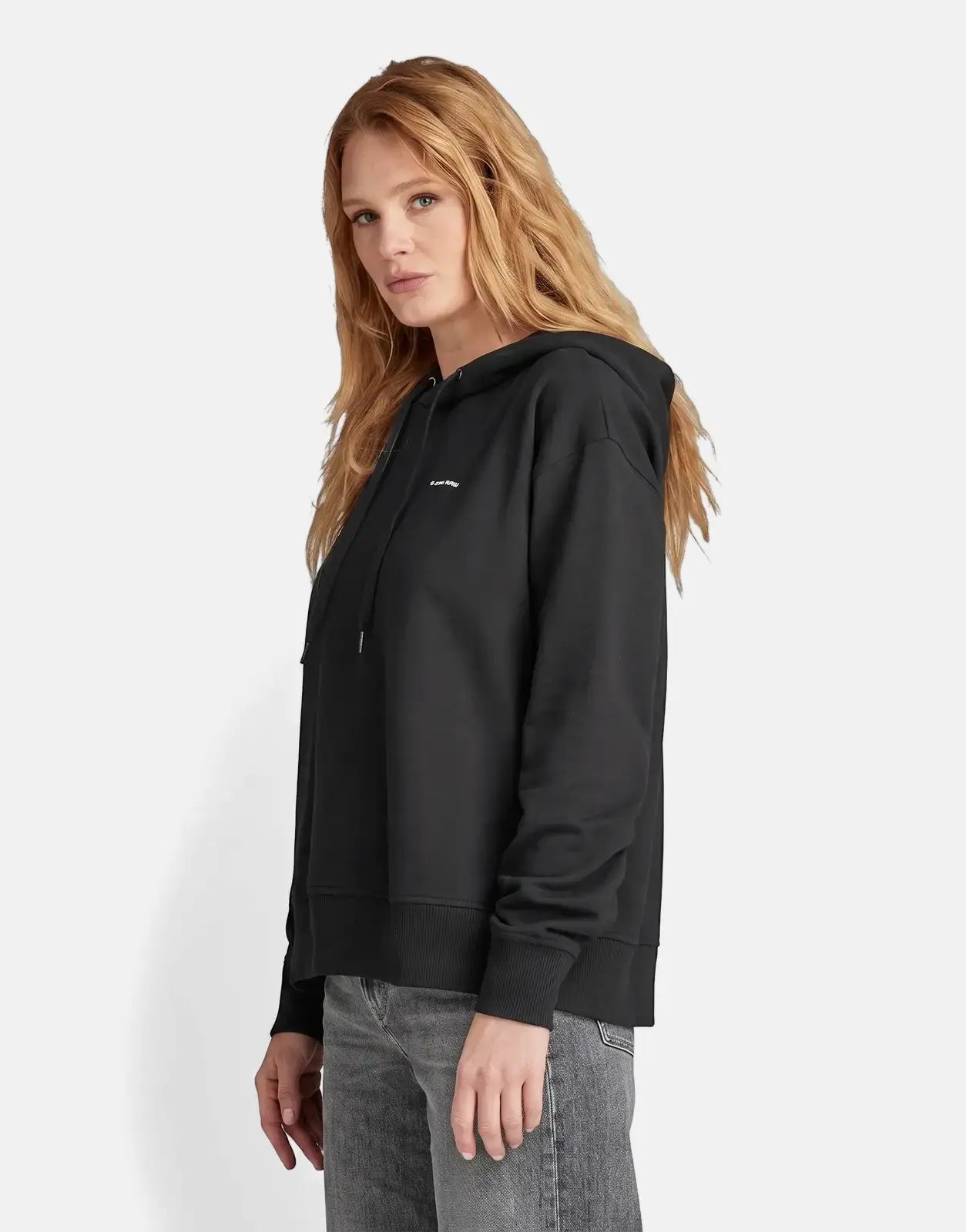 Woman in a black G-Star RAW Graphic Core hoodie, styled with grey jeans, posing in a studio setting.