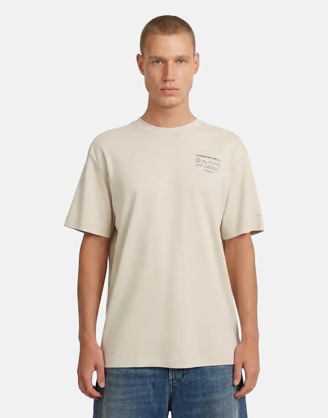 Male model wearing a G-Star RAW Script Whitebait T-Shirt in a neutral beige, paired with blue denim jeans, against a light