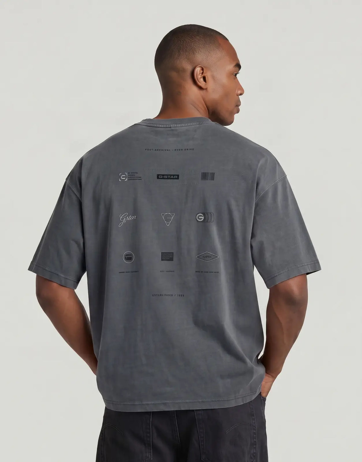 A man models the back of a dark grey G-Star RAW Washed Multi GR Oversized T-Shirt with various graphic prints.