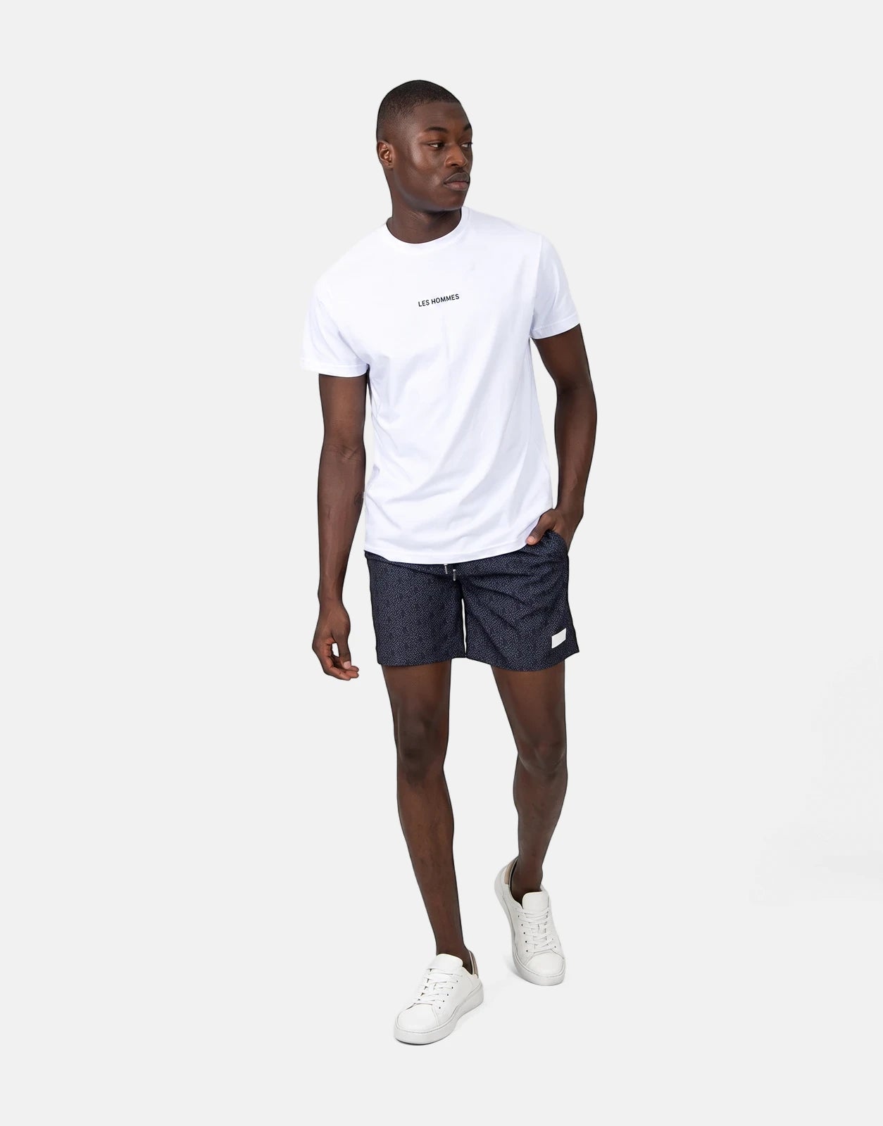 Male model wearing Les Hommes AOP black patterned swim shorts, a white t-shirt, and white sneakers.
