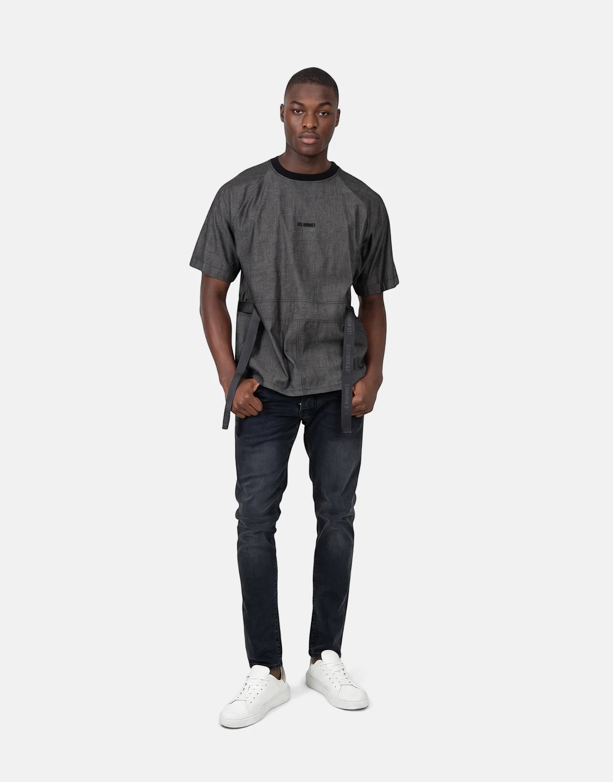 A male model wearing a grey Les Hommes t-shirt with black belt-like straps, black jeans, and white sneakers, standing