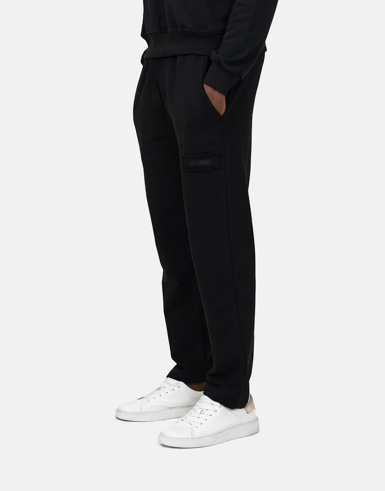 Men's Les Hommes black fleece joggers, seen from a side view, featuring a logo patch and worn with white sneakers.
