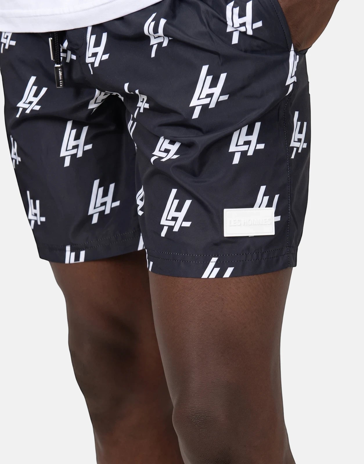 Close-up of Les Hommes black swim shorts with white monogram print and drawstring worn by a model, side view.