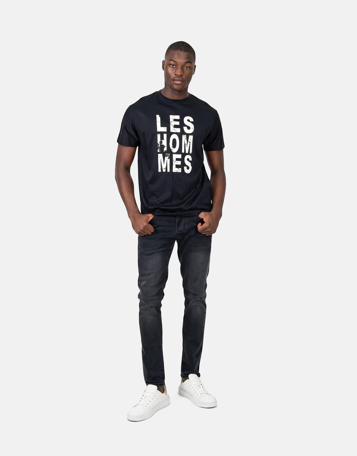 Full body shot of a male model wearing a Les Hommes blue chest graphic t-shirt, dark wash jeans, and white sneakers.