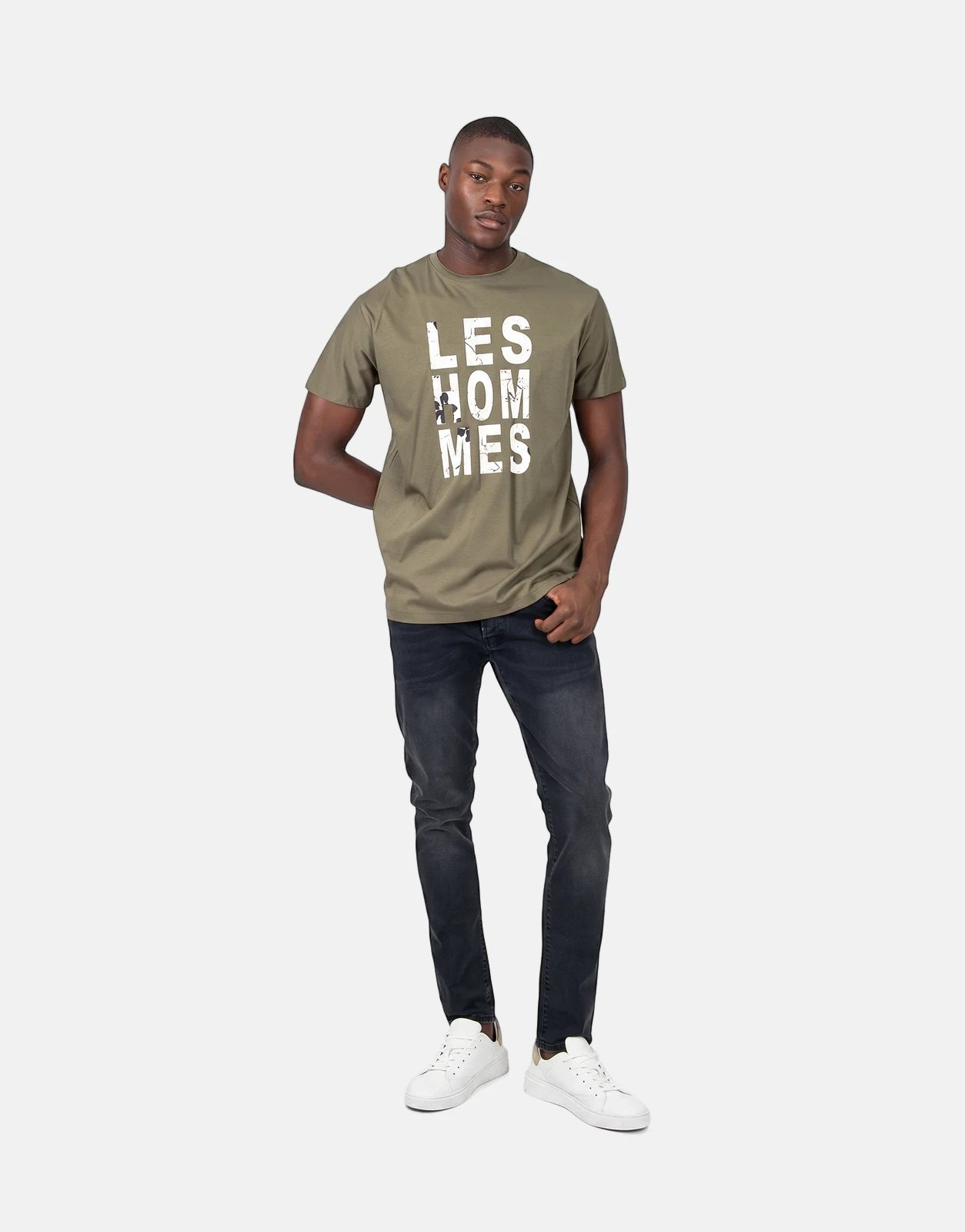 Male model wearing a Les Hommes chest graphic green t-shirt, dark gray jeans, and white sneakers, standing front view.