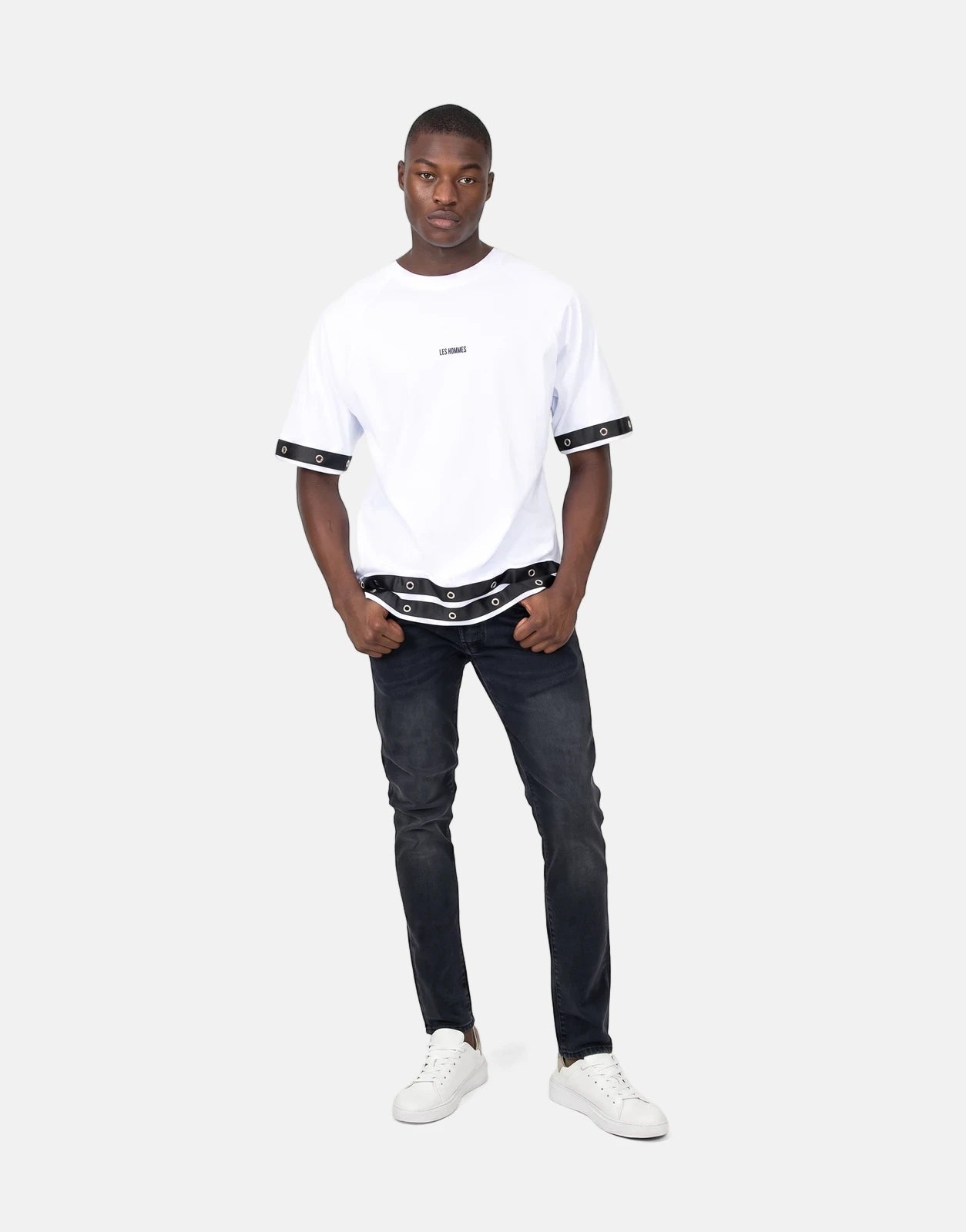 A full shot of a male model wearing a Les Hommes Denim Insert White T-Shirt with black distressed jeans and white sneakers.