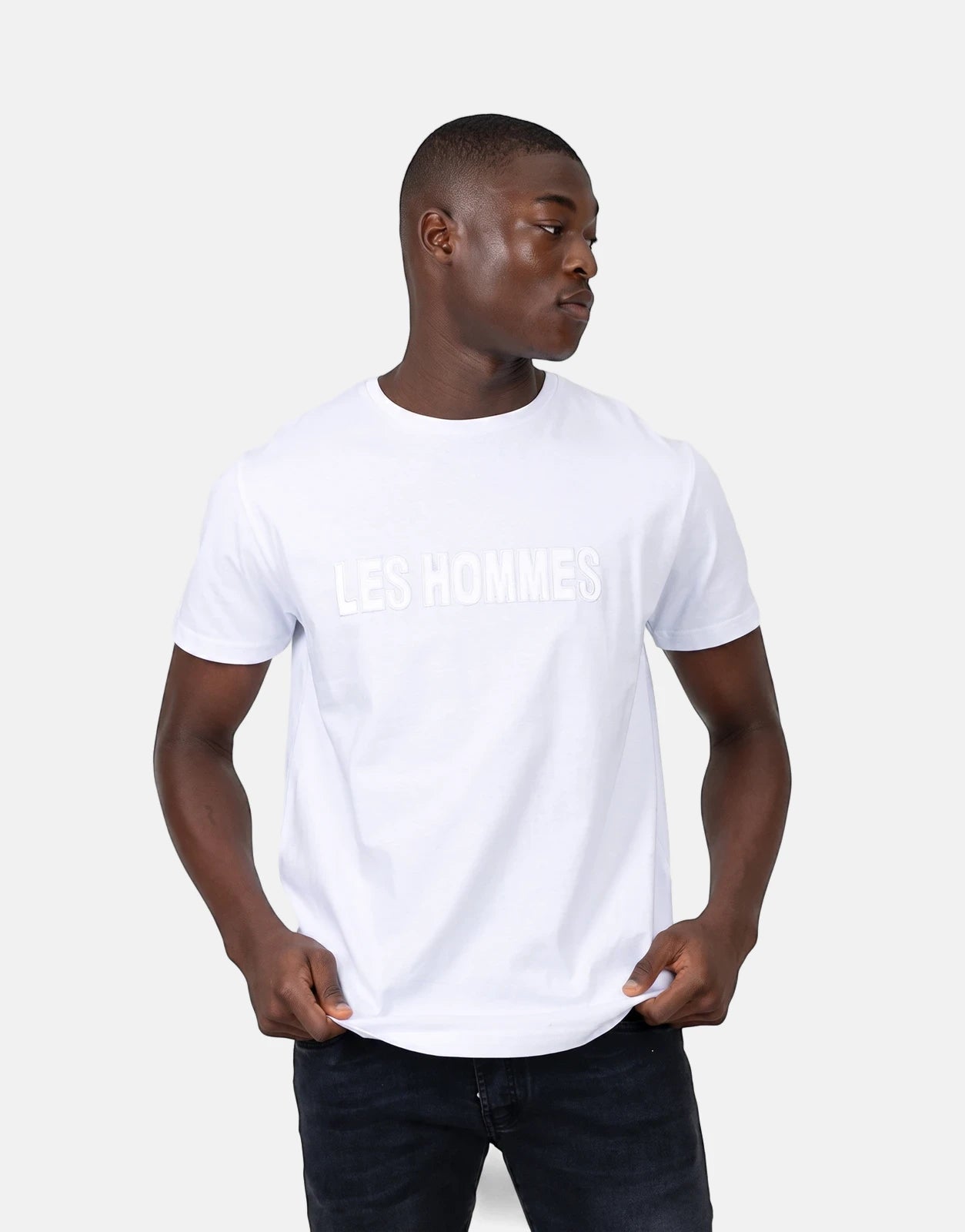 A male model wears a Les Hommes white short-sleeve t-shirt with a glitter logo and dark jeans, looking to the side.