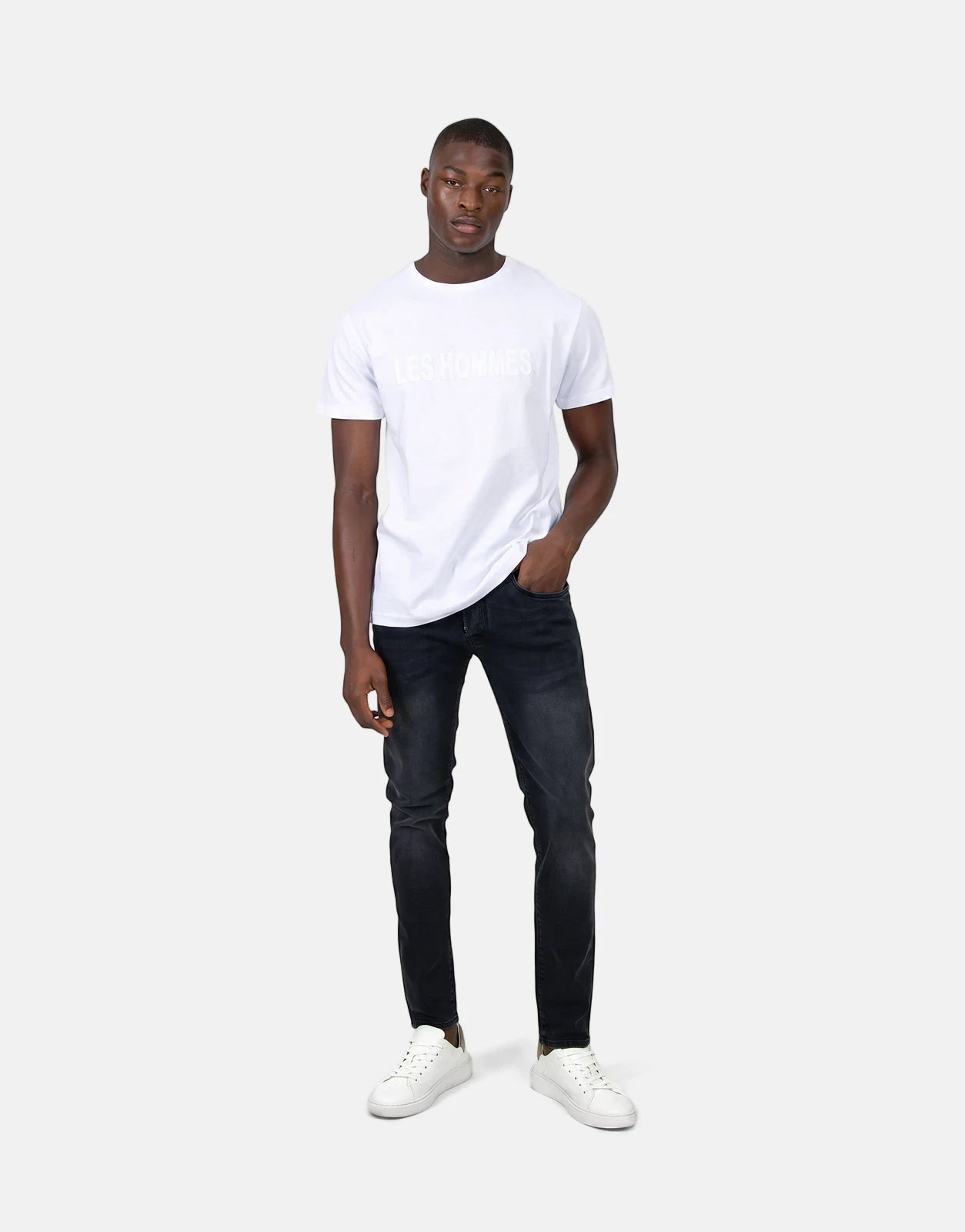 A young man wears a white Les Hommes t-shirt with a glitter logo, black jeans, and white sneakers, posing full-body.