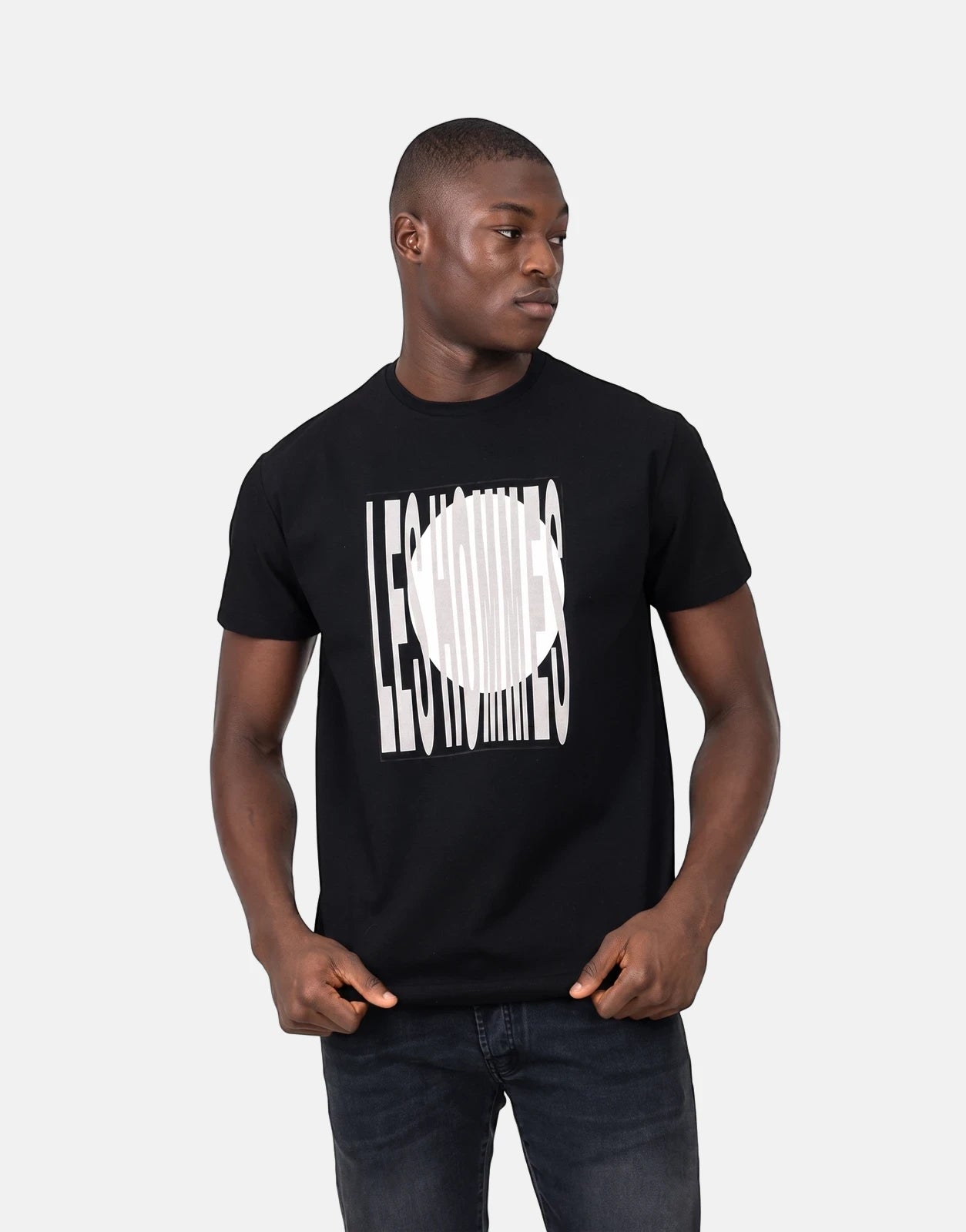 A male model wears a black Les Hommes t-shirt featuring a distinctive white logo print, paired with dark jeans, posed in a