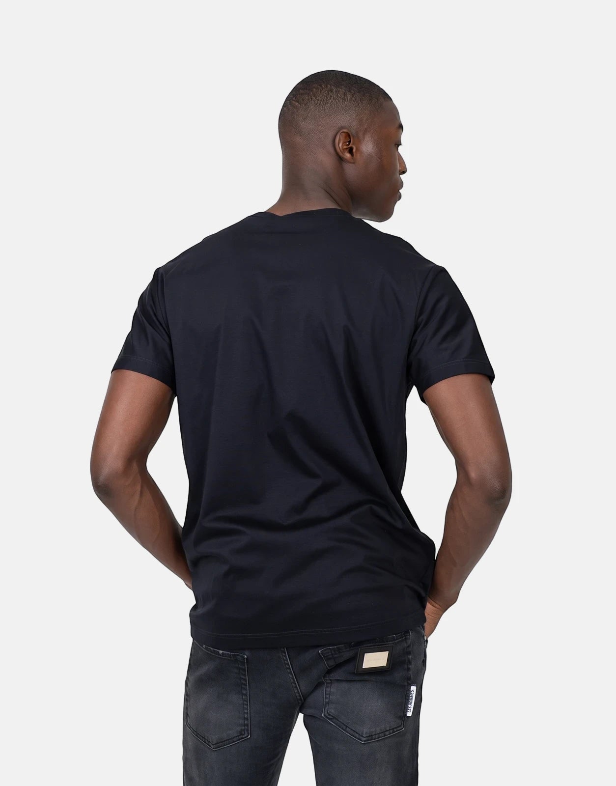 Male model wearing a black Les Hommes t-shirt and dark gray jeans, viewed from the back with hands in pockets.