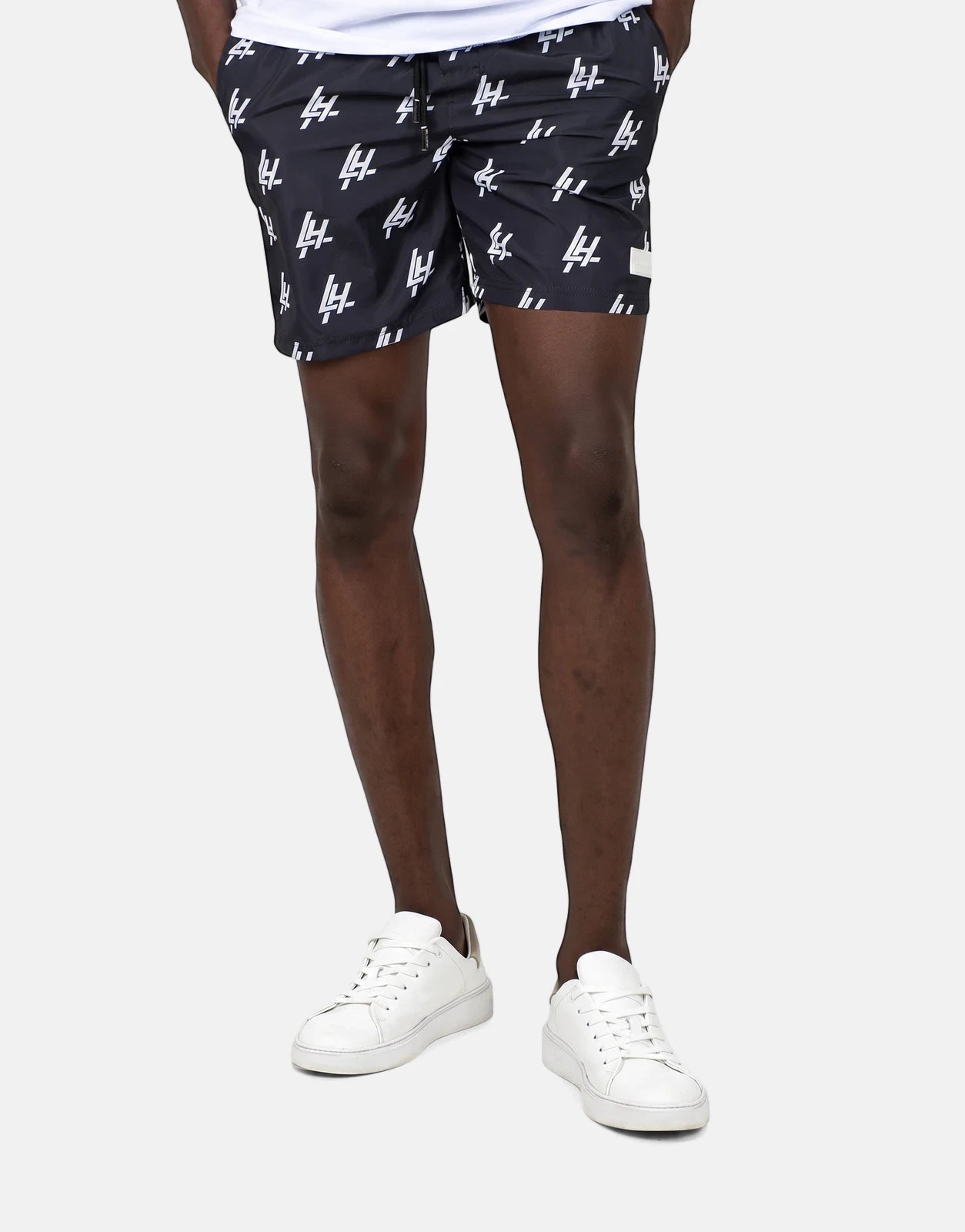 Men's Les Hommes black swim shorts with white repeating print, shown on a model from the mid-thigh down, paired with white