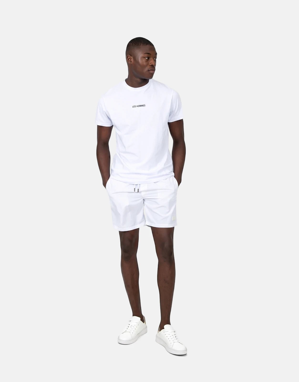 Full-body shot of a male model wearing Les Hommes white swim shorts, a white t-shirt, and white sneakers.
