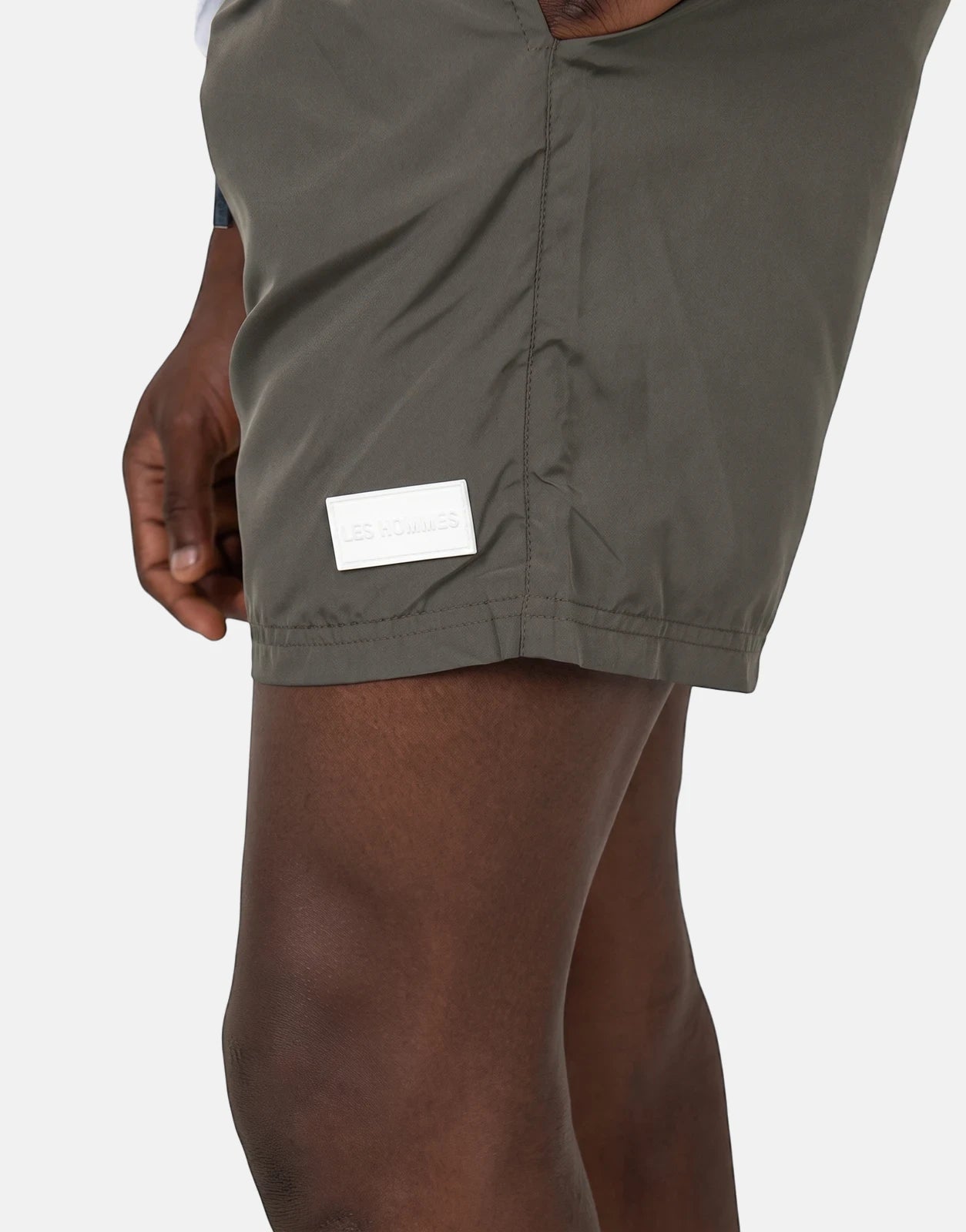 Close-up of Les Hommes olive green swim shorts worn by a model, featuring a white rectangular logo patch on the thigh.