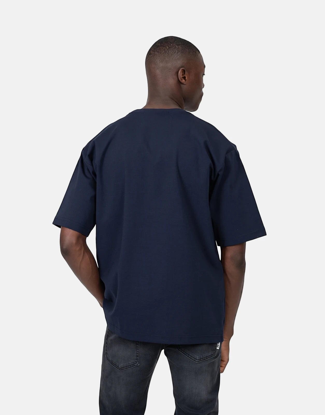 Rear view of a male model wearing a dark blue Les Hommes oversized t-shirt and dark wash jeans in a studio setting.