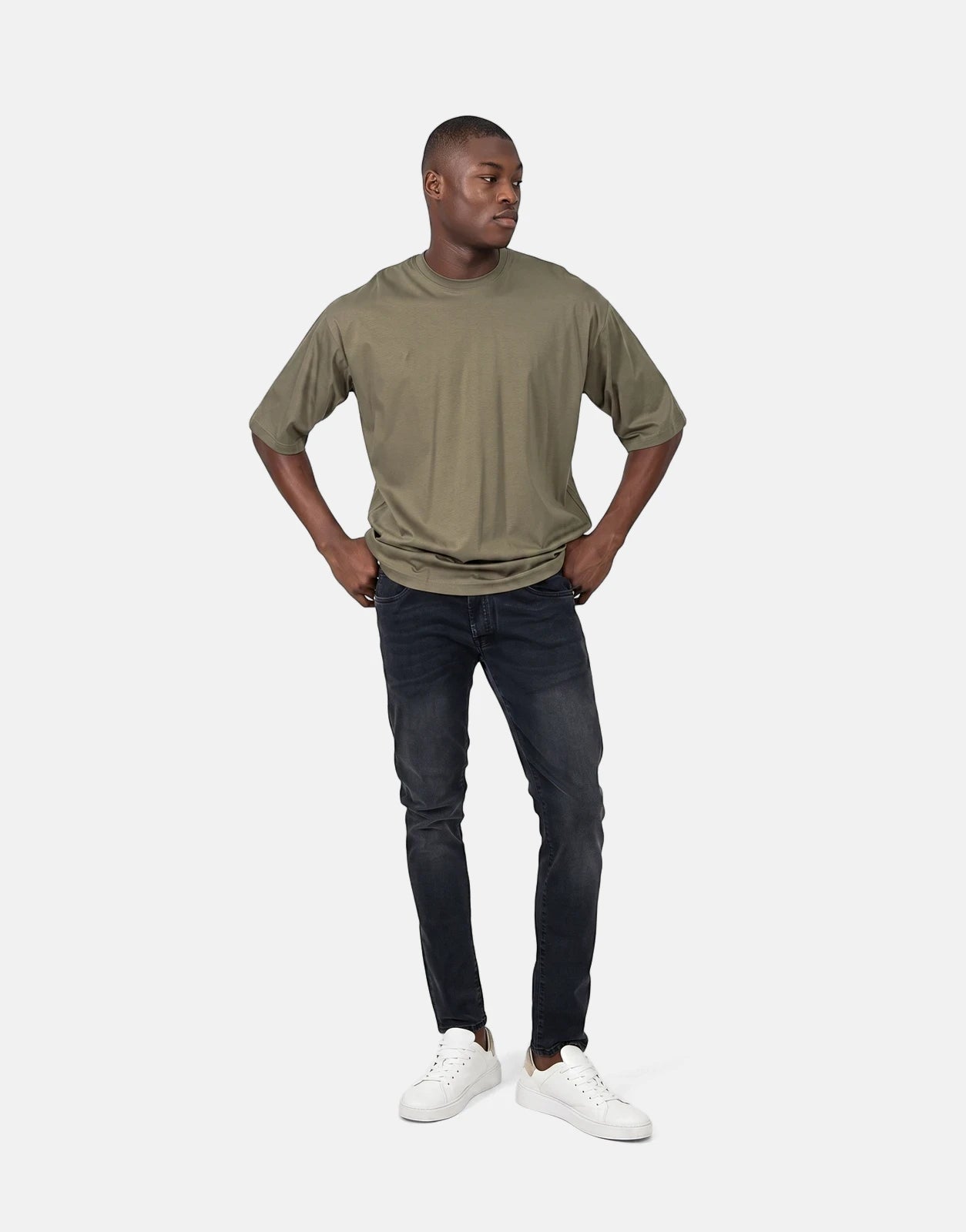 Young man models a Les Hommes oversized olive green short-sleeve t-shirt, paired with dark wash slim-fit jeans and white