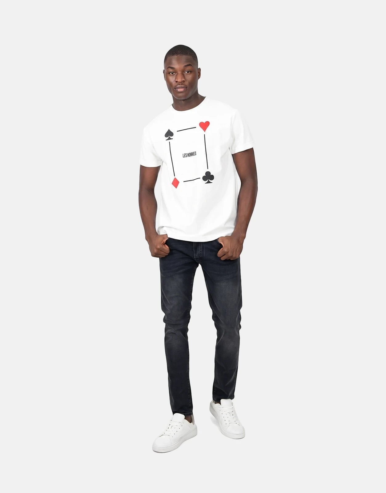 A male model wearing a Les Hommes Poker Print White T-Shirt, black jeans, and white sneakers, posed full-body.