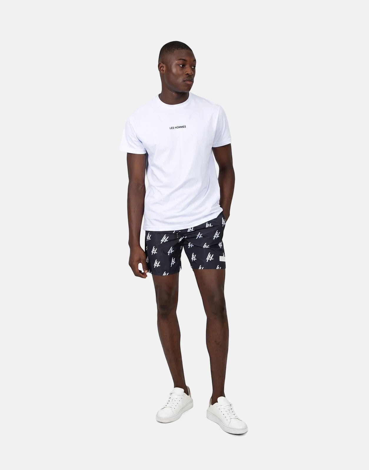 Full body shot of a male model wearing Les Hommes Print Black Swim Shorts, a white t-shirt, and white sneakers.