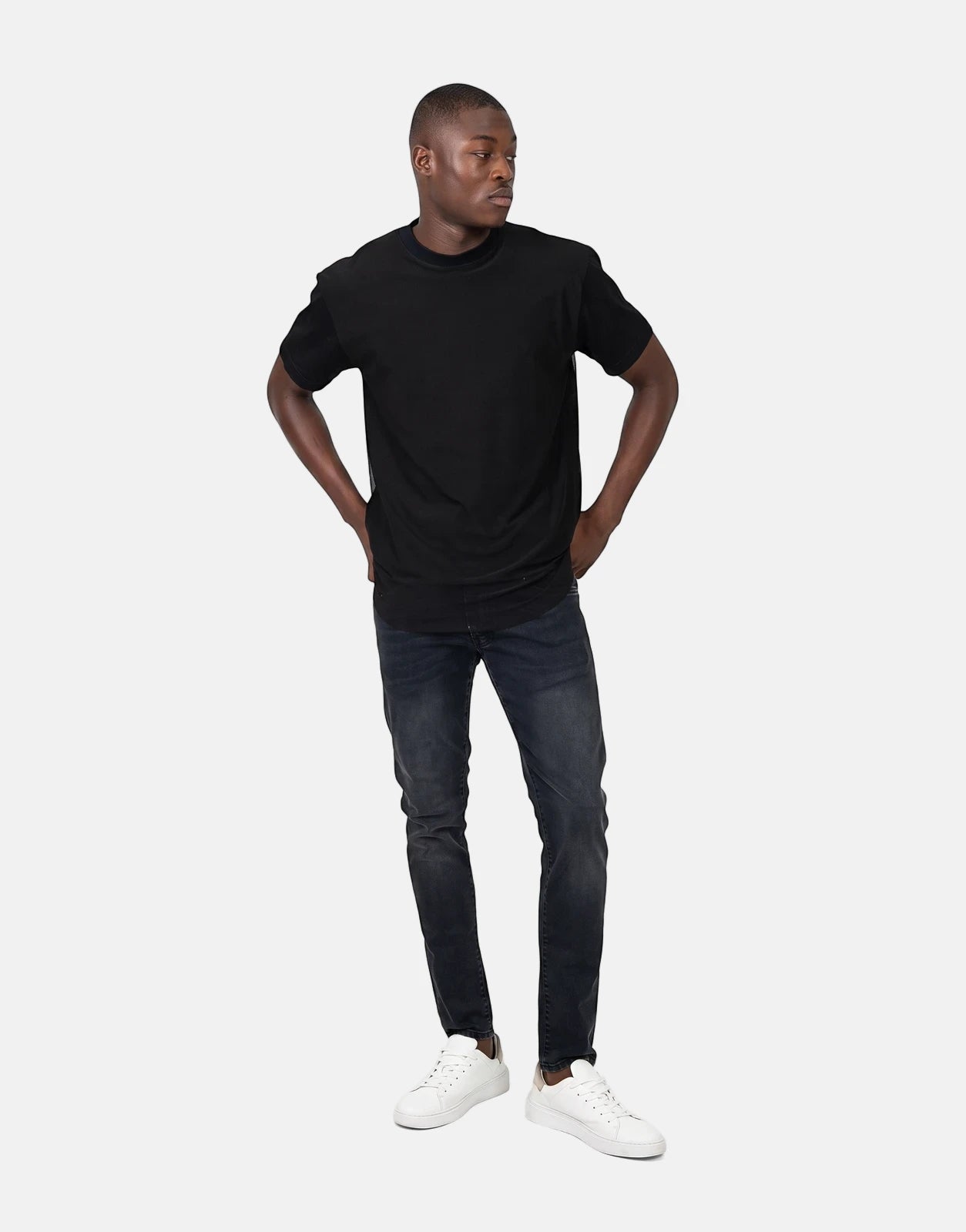 A male model wears a Les Hommes Tulle black short-sleeve t-shirt, dark jeans, and white sneakers.