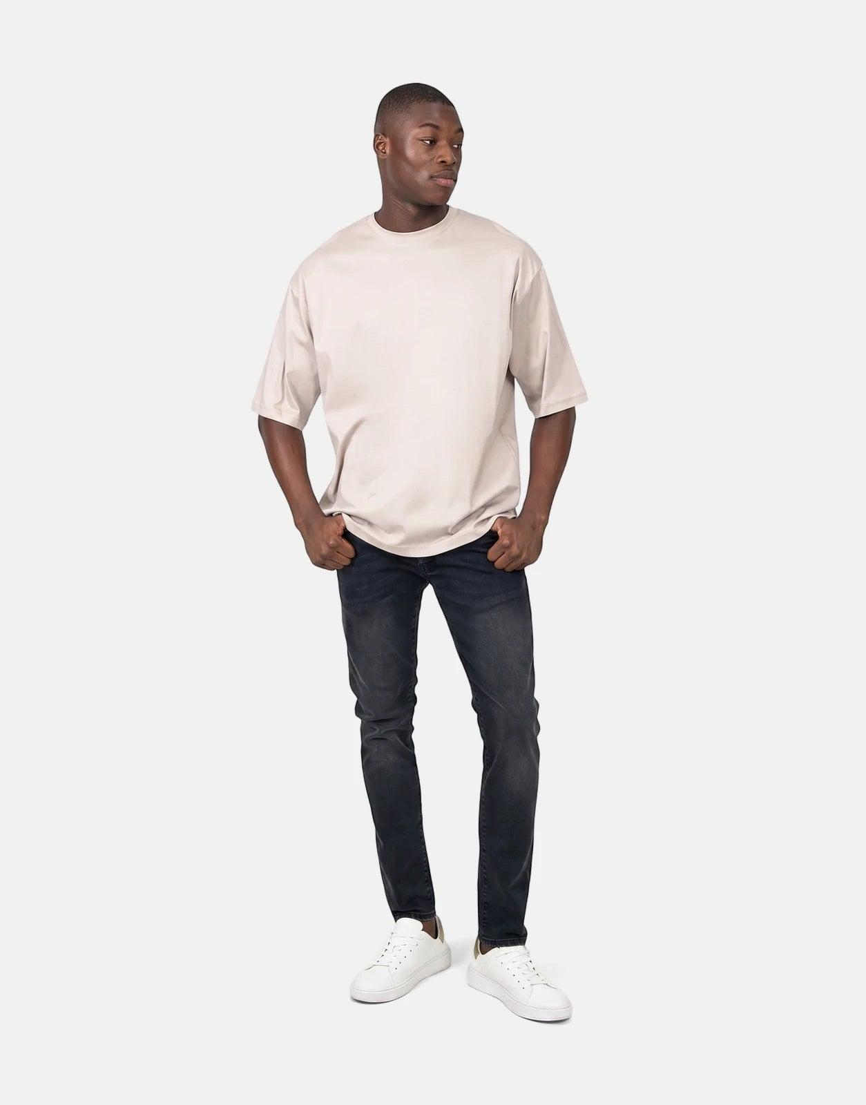 A young man stands, full body, wearing a plain beige oversized crew-neck t-shirt, dark grey skinny jeans, and white sneakers.