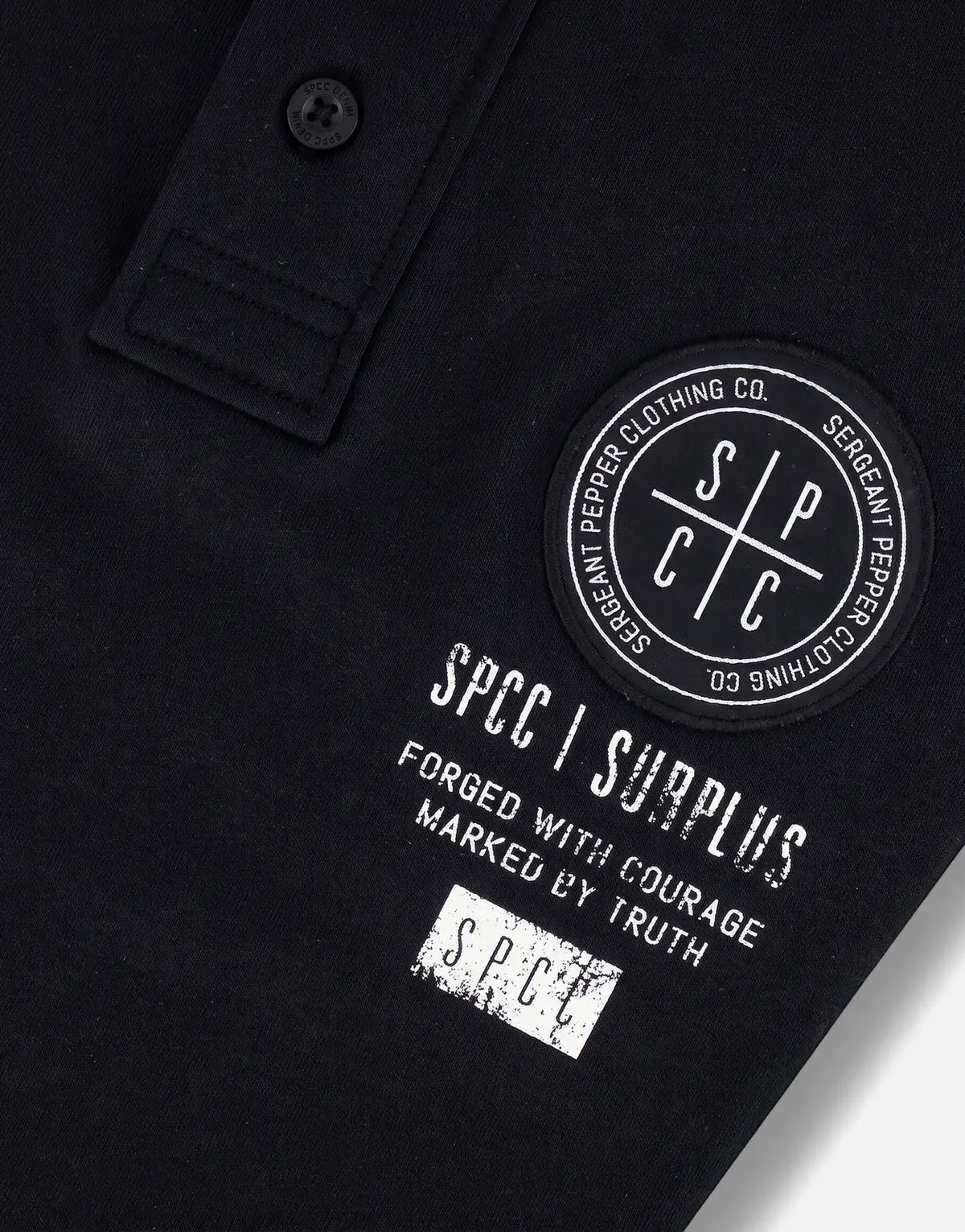 Close-up of a black SPCC Roarke Golfer polo shirt, featuring the embroidered circular brand patch and distressed text prints.