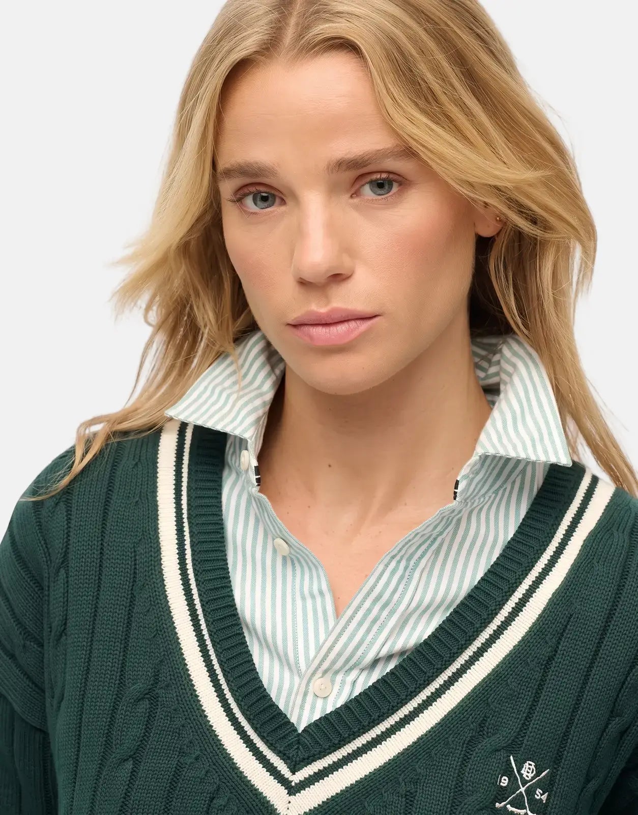 A blonde woman wears a Superdry Cricket Club Enamel Green Jumper over a striped shirt, showcasing the cable knit texture.