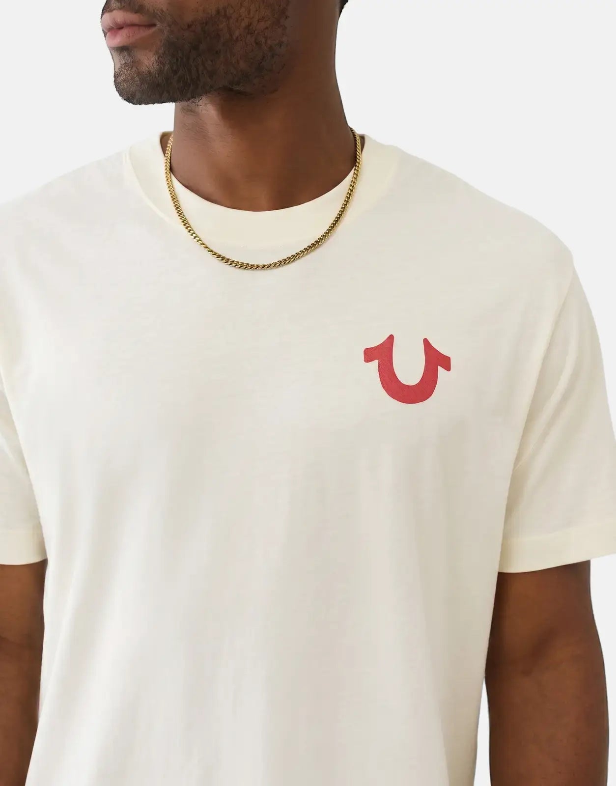 Close-up of a man wearing an off-white True Religion Number One SRS T-shirt with a red horseshoe logo and a gold chain.
