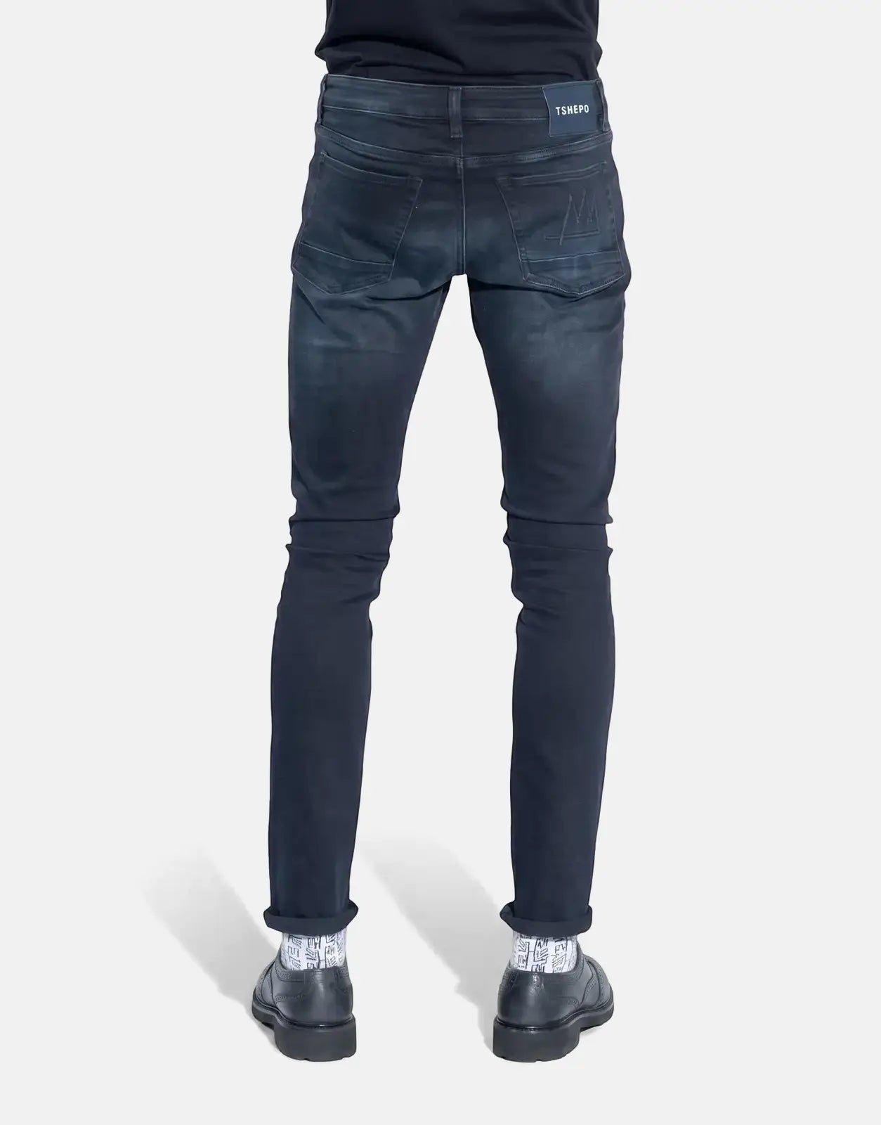 Back view of men's TSHEPO Takalani slim fit dark wash jeans, worn with patterned socks and black shoes.