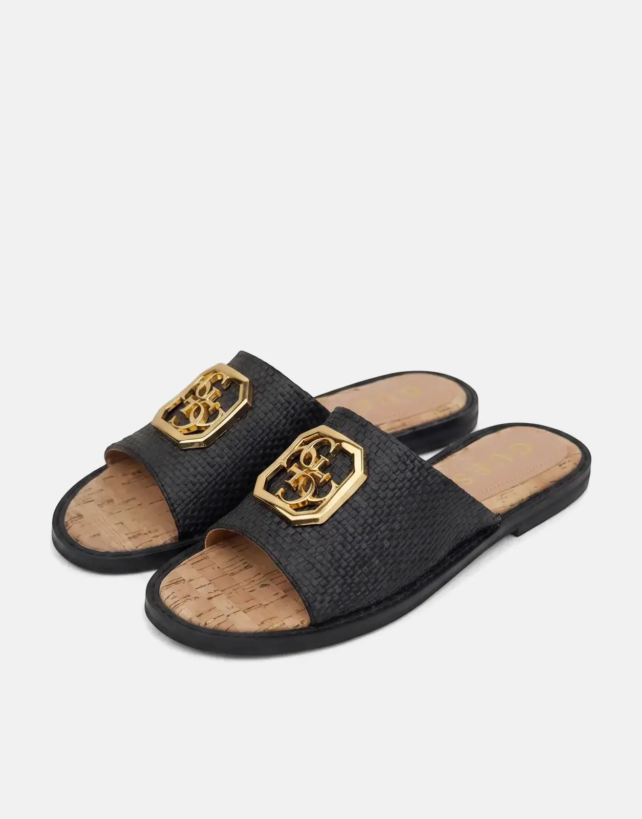 Pair of women's black woven slides with gold Guess logo hardware and cork-patterned footbeds, angled view.