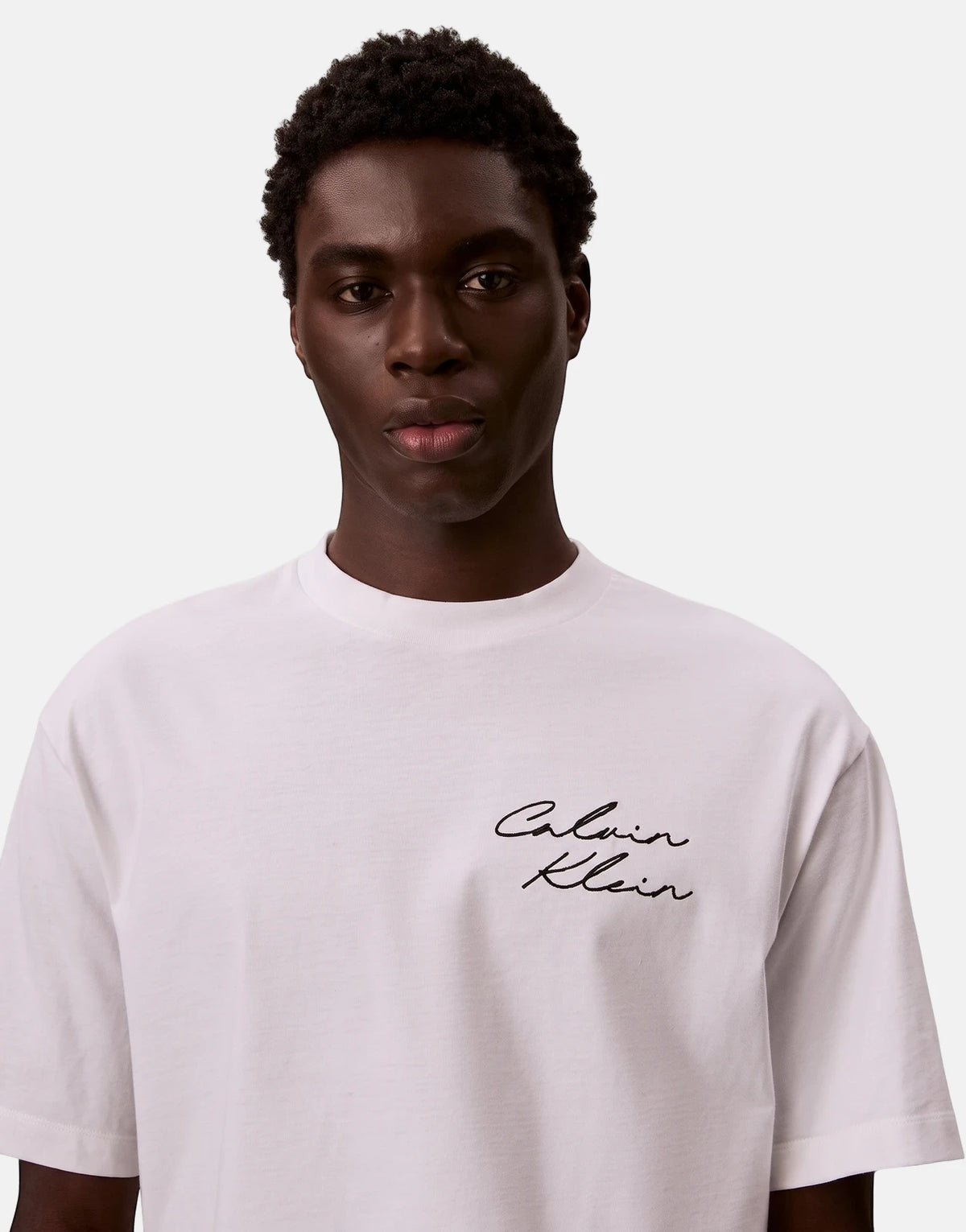 A dark-skinned male model wears a white Calvin Klein t-shirt with a black script logo on the chest, looking forward.