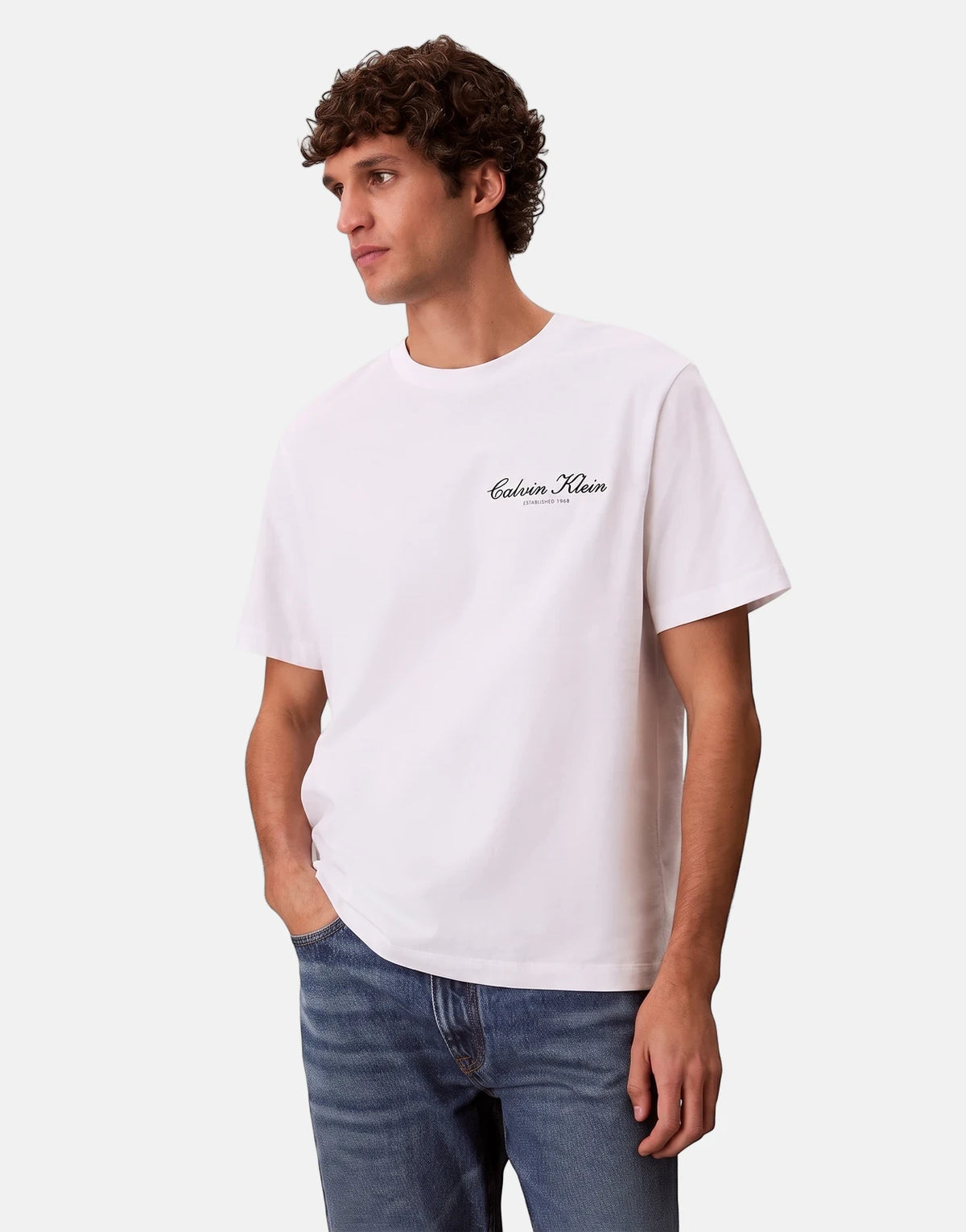 Male model wearing a white Calvin Klein graphic t-shirt and blue jeans, standing casually with hand in pocket.