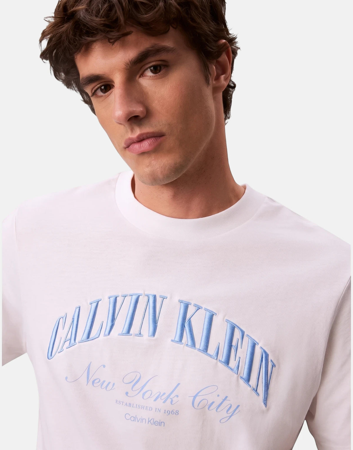 A man wears a white Calvin Klein 20s March graphic seasonal t-shirt with blue embroidered 'New York City' logo.