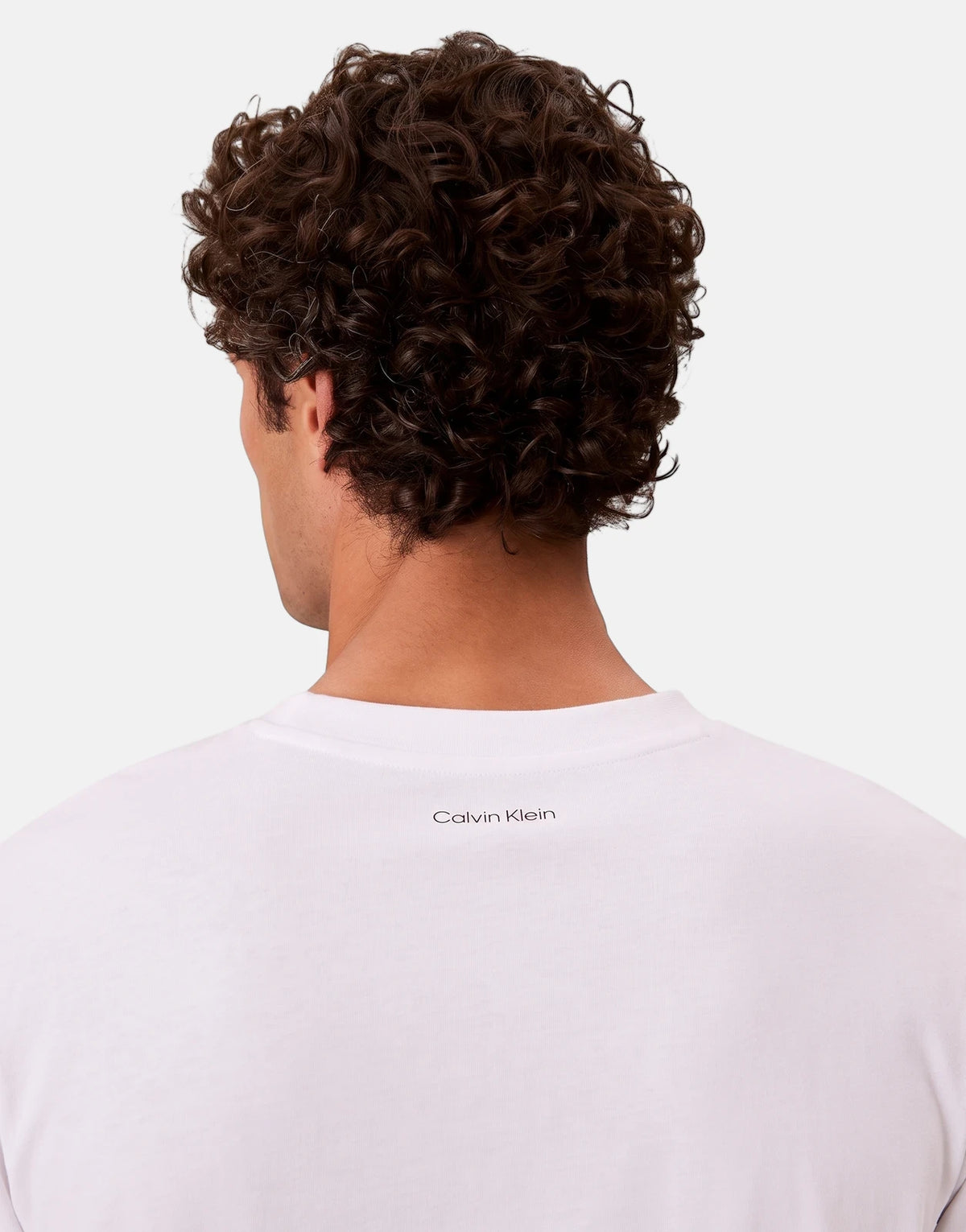 Back view of a male model with curly hair wearing a white Calvin Klein 20s March Graphic Seasonal T-Shirt with the logo