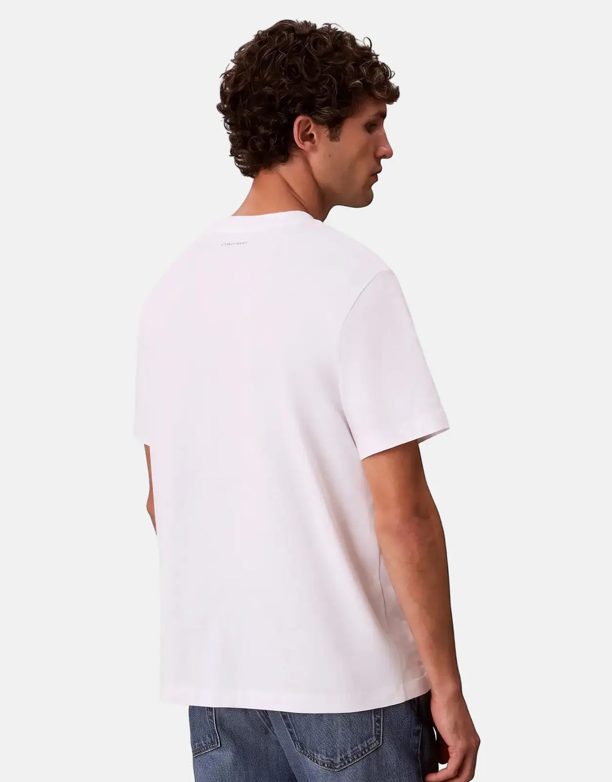 Male model from behind, wearing a Calvin Klein 20s March Graphic Seasonal white t-shirt and blue jeans, with curly brown