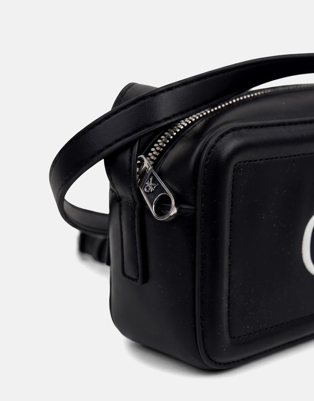 Black Calvin Klein camera bag made of faux leather, featuring a prominent silver zipper with a CK logo pull. Close-up view.
