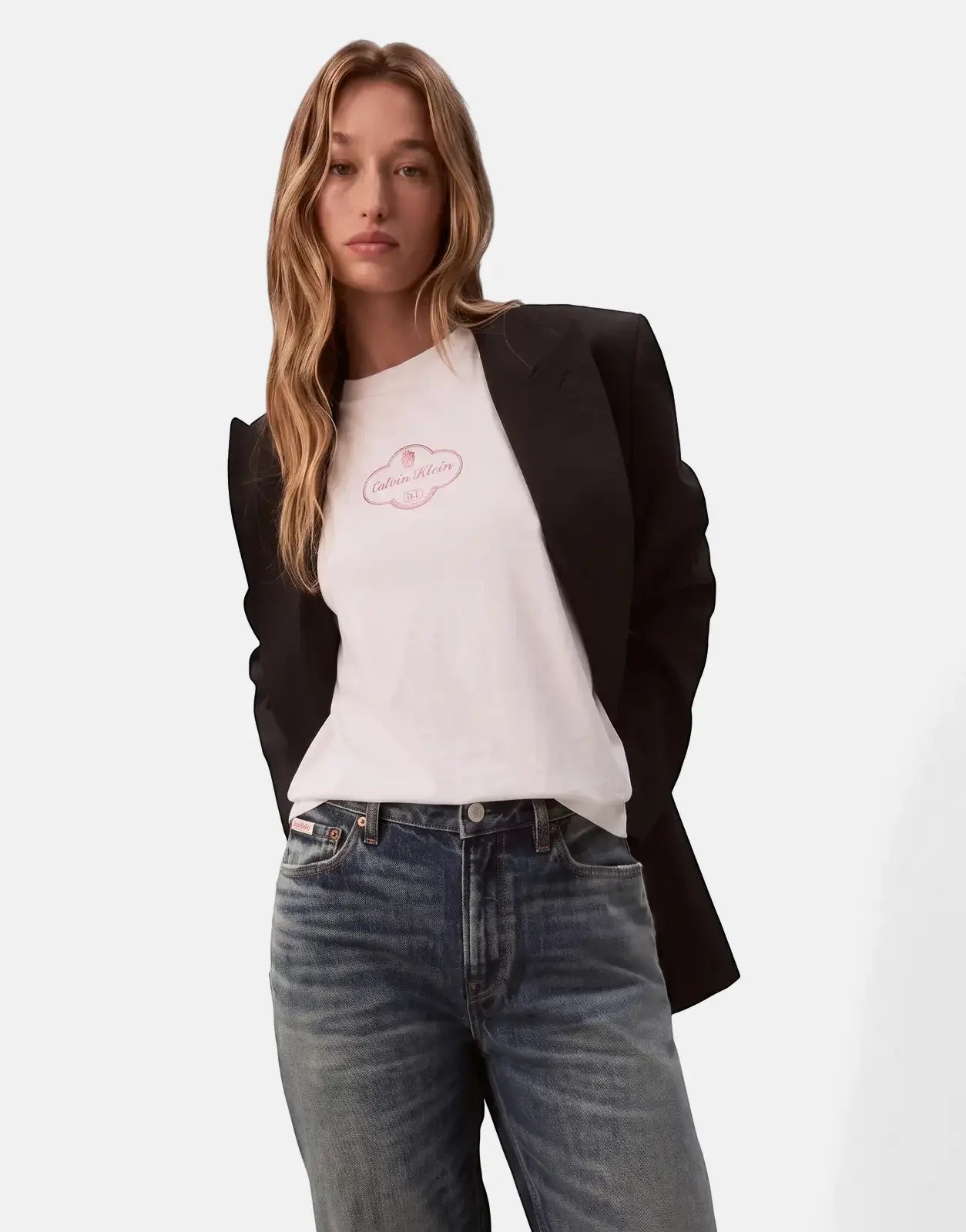Female model in a white Calvin Klein Boxy Cocktail Logo T-shirt, black blazer, and blue jeans, posing.