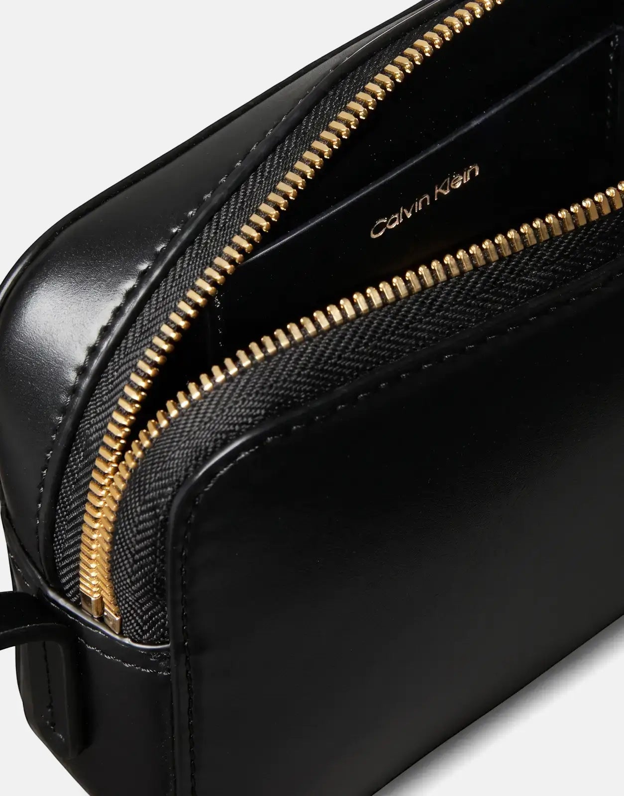Close-up of a black Calvin Klein camera bag with an open gold zipper compartment, revealing the brand logo inside.
