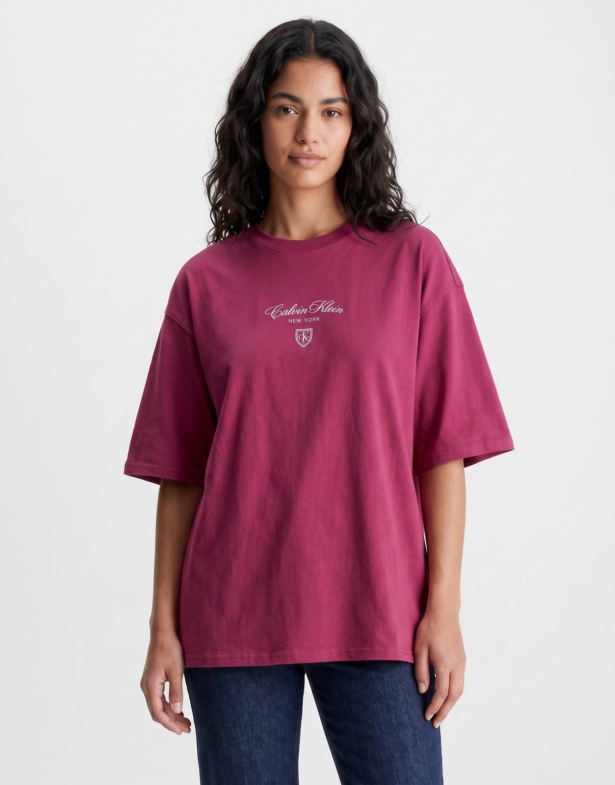 A woman models a Calvin Klein CK NY oversized berry t-shirt, featuring a white logo on the chest, paired with dark jeans.