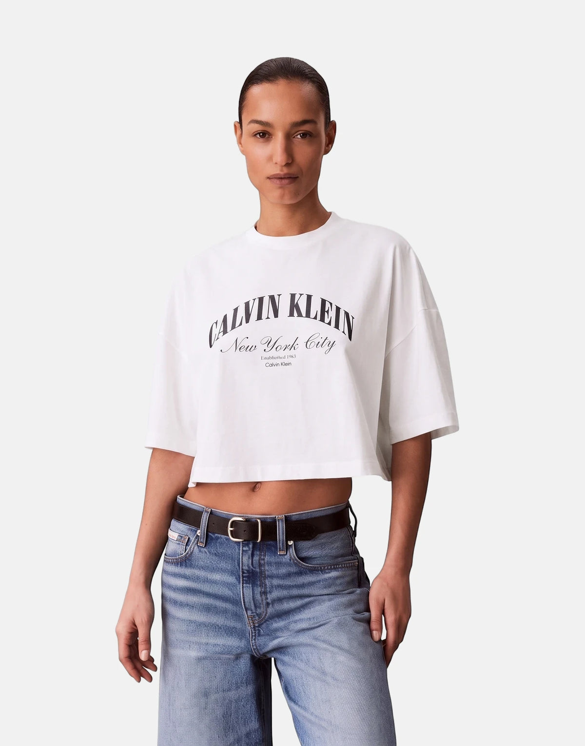 A female model wears a white Calvin Klein Cropped Boxy Heritage Crest Logo T-Shirt paired with blue denim jeans and a black